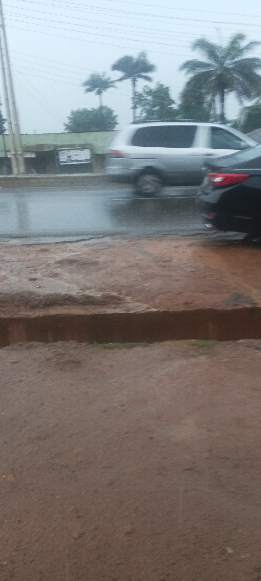 The image depicts a street scene in Akwanga, Nigeria, during a rainstorm. The foreground shows a dirt sidewalk with a small drop-off. In the background, there is a wet asphalt road with two cars: a silver minivan moving to the left and a parked black sedan. Palm trees are visible beyond the road. The sky is overcast and grey, indicating the rain.