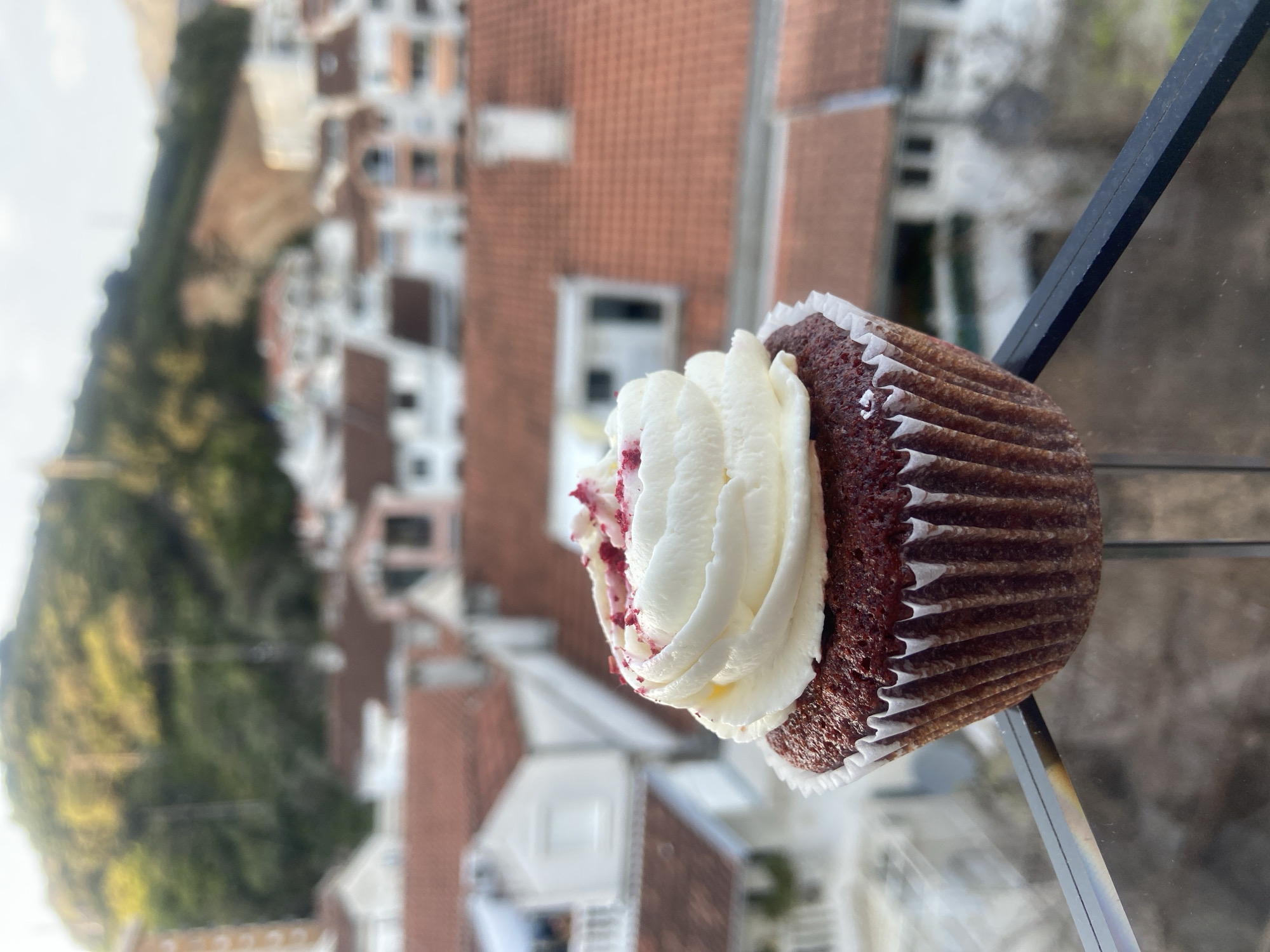 A cupcake, possibly red velvet with white frosting and red sprinkles, sits in the lower right quadrant of the frame on a shiny surface, potentially glass or plexiglass. In the background, blurred, is a cityscape, possibly Budva, Montenegro, based on the architecture: white buildings with red tile roofs. A green vegetated area is present in the background on the left. A black vertical object is partially visible to the right of the cupcake. The image has a shallow depth of field, focusing mainly on the cupcake.