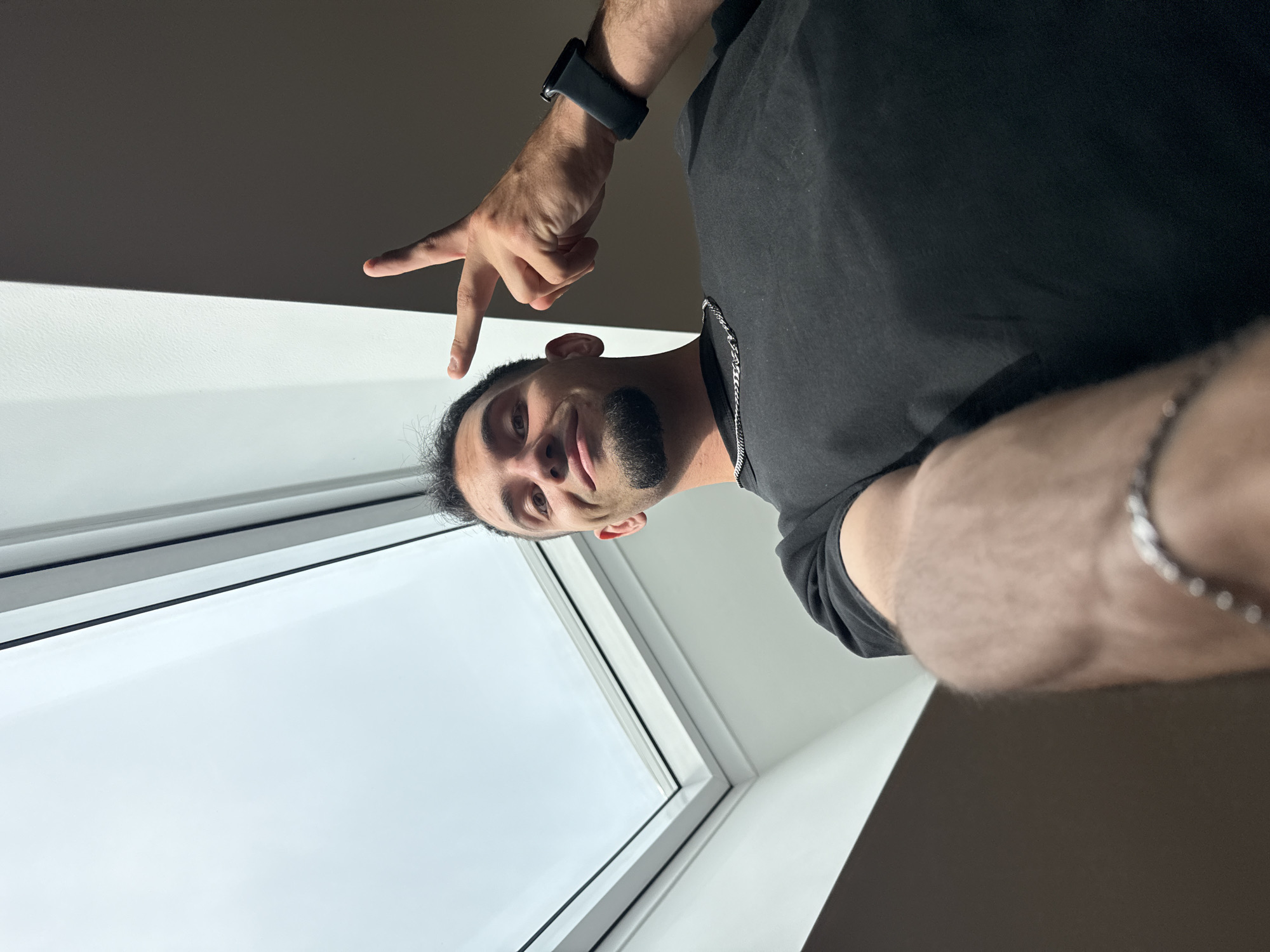 A man is looking up at a skylight, holding up two fingers in a peace sign. He is wearing a black t-shirt and a silver chain. He has a goatee and is wearing a watch on his left wrist. The room is dimly lit, with the skylight providing most of the illumination. The overall scene is consistent with an indoor setting, potentially a home or an office, in Garmisch-Partenkirchen, Germany.