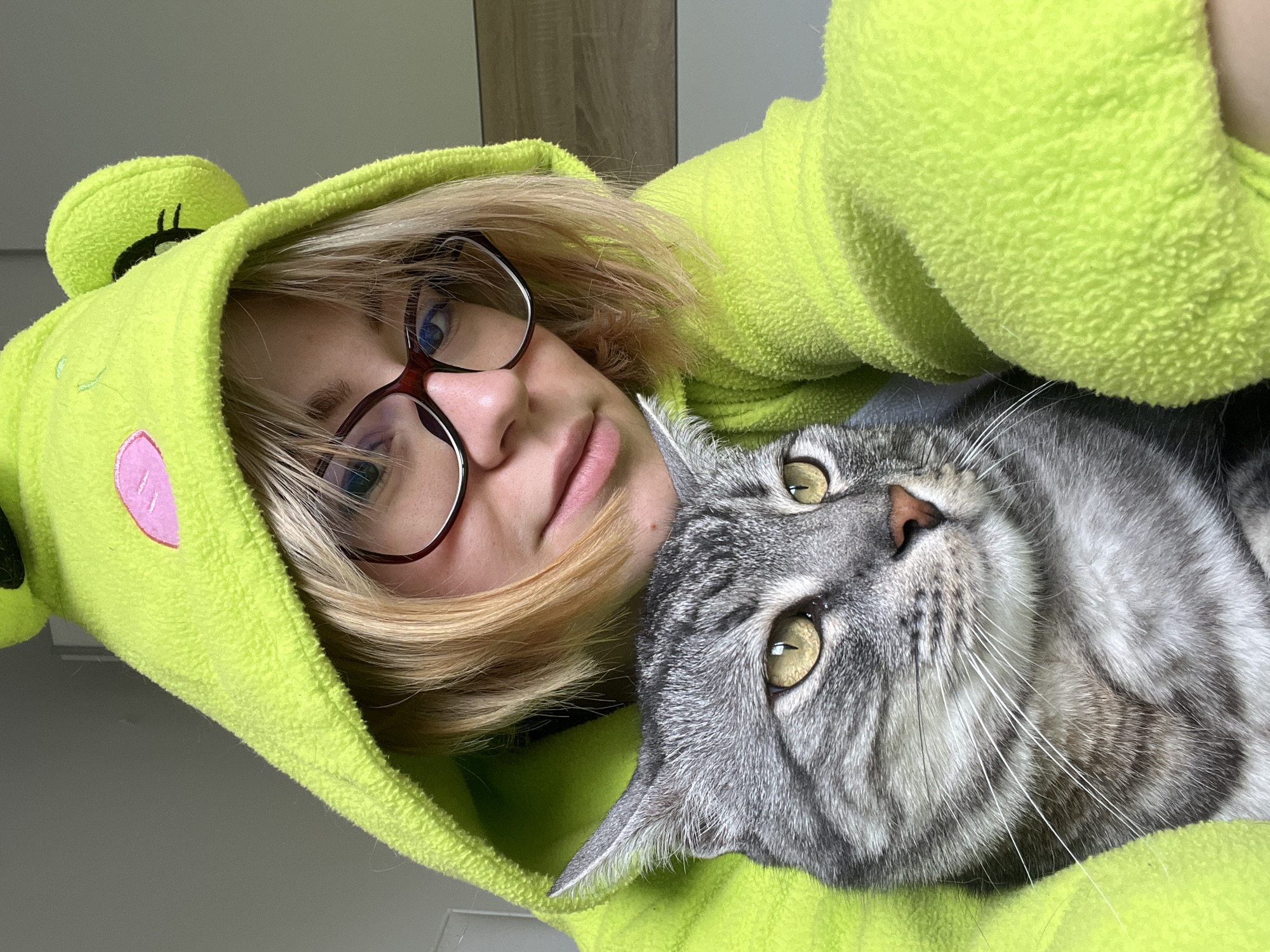 A close-up, slightly angled photograph shows a person and a cat. The person, presumably female, is wearing glasses and a bright green frog-themed hooded garment. The cat, a silver tabby, is nestled closely to the person. The background is out of focus and appears to be an interior space with light-colored walls and possibly wood paneling in the upper background. It is impossible to determine if this photo was taken in Boreti, Montenegro based on the information given. There are no other people, subjects, objects, or actions in the immediate view. The main interaction is the close proximity of the person and the cat. The environment is nondescript indoors.