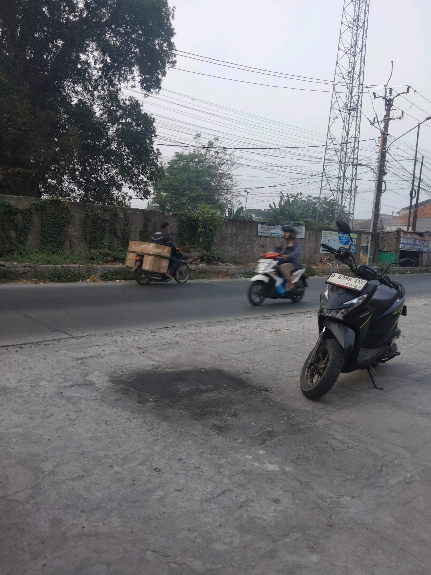 In Pisangan Jaya, Indonesia, on an overcast day, a street scene unfolds. A dark-colored scooter with a license plate reading "A 6386 XKC" is parked on a cracked concrete surface in the foreground. The scooter faces left, its front wheel turned slightly towards the camera. In the background, a two-lane asphalt road is visible. On the road, a person on a black motorcycle is transporting several large cardboard boxes stacked on the rear seat and extending upwards. Further down the road, another person rides a white scooter. The roadside is lined with a low concrete wall covered in green ivy, beyond which are trees and a tall lattice tower, characteristic of telecommunication infrastructure. Electrical wires crisscross the sky, typical of Indonesian urban environments. The overall impression is one of everyday commerce and infrastructure in a bustling Indonesian town.