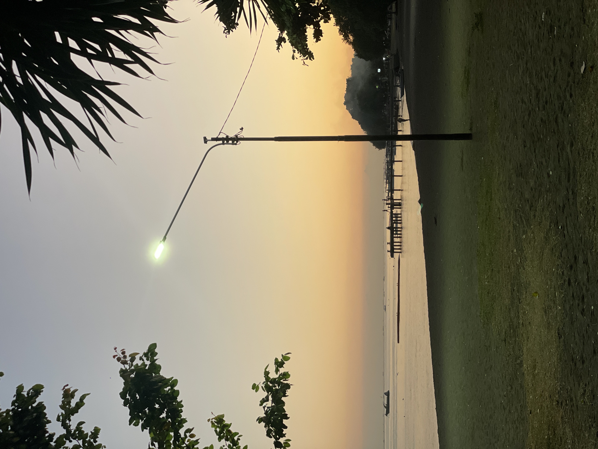 The image depicts a beach scene at dusk in Buleleng, Indonesia. The sky exhibits a gradient of soft yellows and oranges, transitioning to a pale blue overhead, indicative of sunset or sunrise. In the foreground, a dark sandy beach with patches of sparse green grass occupies the lower portion of the frame. A tall, dark lamppost stands prominently in the mid-right, its light illuminated and casting a greenish glow. The middle ground features a body of calm water, likely the sea, with several small boats and a pier extending into the water. Silhouettes of people can be seen on the pier, some appearing to be fishing or strolling. The far background is characterized by a treeline and a small hill, partially obscured by mist or smoke. Tropical foliage frames the top left and right corners of the image, with palm fronds visible on the right. The overall atmosphere is serene and tranquil, capturing a moment of twilight by the coast.