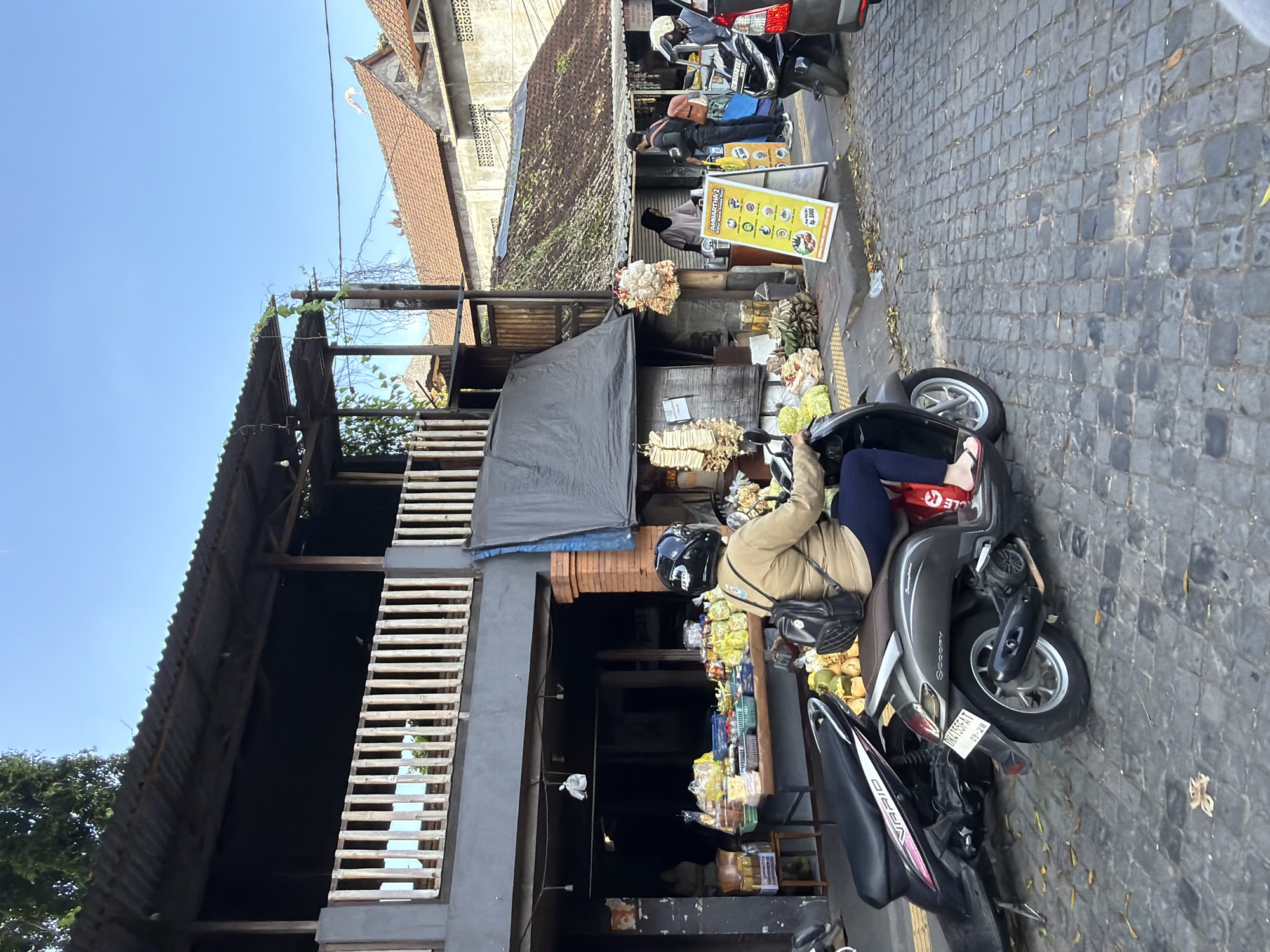 The image depicts a street scene in Ubud District, Indonesia. A woman wearing a beige jacket, dark blue pants, and a black helmet is riding a dark gray scooter on a cobblestone street. She has a black backpack on. Two other scooters are parked nearby. In the background, there is a market stall with various goods displayed, including fruits and packaged items. A yellow sign with Indonesian text is visible in front of a stall where two individuals are standing. The architecture features traditional Indonesian elements with tiled roofs and wooden structures. The sky is clear and blue, indicating a sunny day.