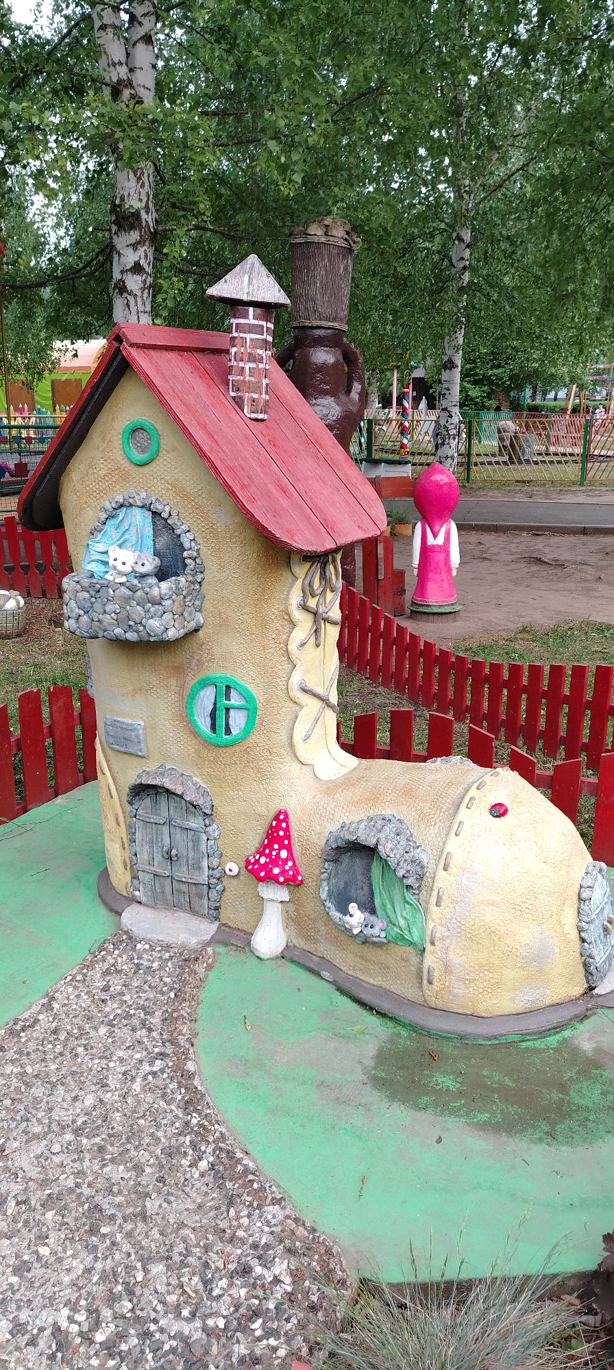 The image shows a whimsical, oversized boot-shaped house in a public space. The "boot-house" has a red roof, a stone pathway leading to a small, grey door, and windows with small animal figures looking out. It is painted in tan and adorned with details like a mushroom and stone accents. A red picket fence surrounds the house. In the background are trees and additional playground or park elements, including a brown figure that seems to be made of stacked pots and a pink statue. The scene appears to be located in a park or playground, possibly in the city of Chaikovsky, Russia, based on the provided context.