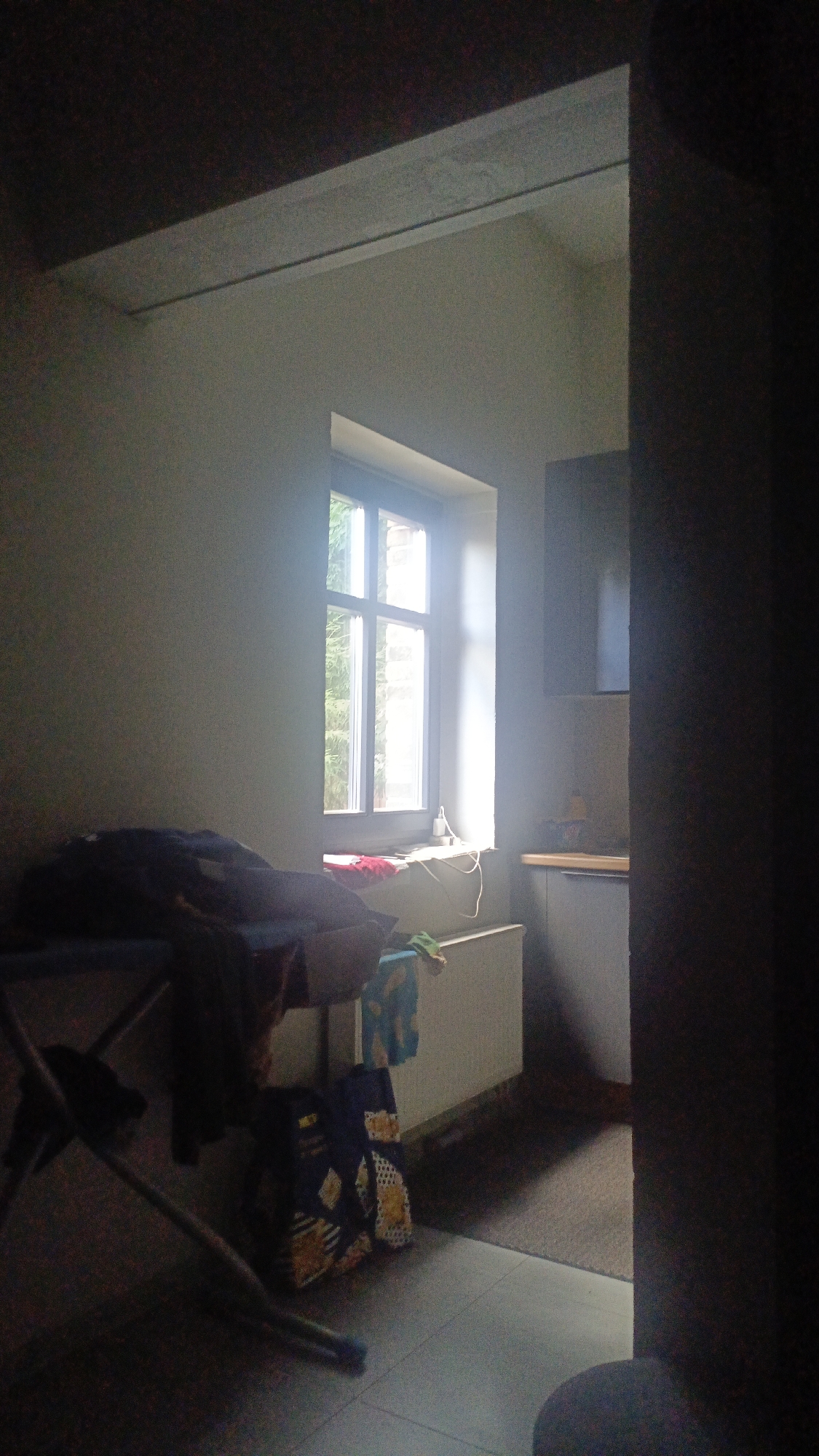 The image depicts a dimly lit interior scene. A prominent window with multiple panes is visible, allowing natural light to enter the room. Outside the window, greenery and the facade of a brick building can be seen. On the windowsill, a red cloth or rug is present, along with some wires. In the foreground, to the left, a clothes rack with dark clothing draped over it is situated. Next to the rack, a blue and yellow patterned bag or backpack rests on the floor. A white radiator is positioned below the window. To the right of the window, a section of kitchen cabinetry with a countertop is visible, suggesting a domestic setting. The overall environment appears to be a room in Nikolino, Russia.