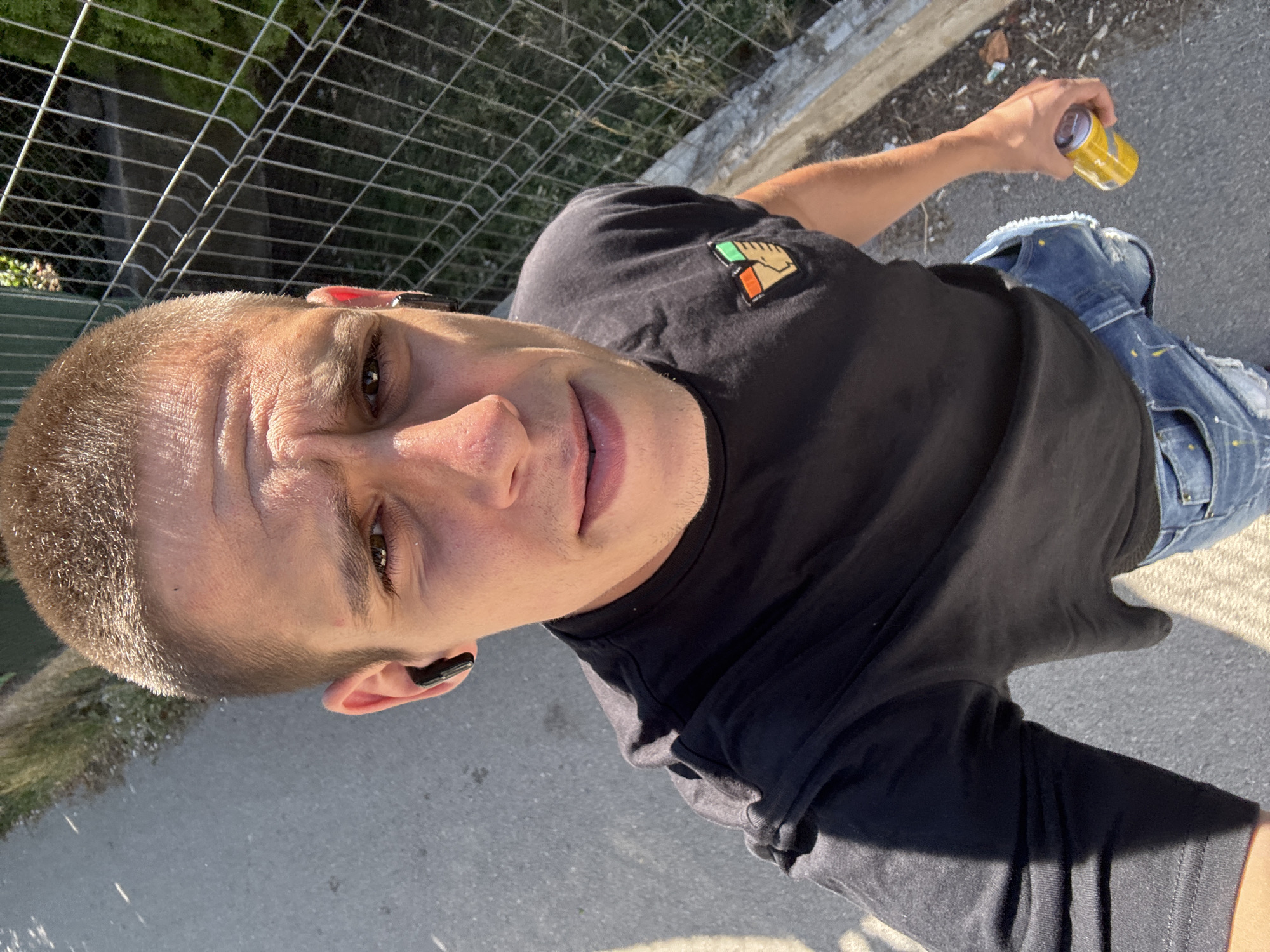A man with a buzz cut and a black t-shirt is standing outdoors. He is wearing a black earbud in his left ear and blue jeans. He is holding a yellow can in his right hand. Behind him, a chain-link fence separates the pavement from an area with grass and greenery. The lighting suggests it is daytime, possibly a sunny afternoon. This image was taken on May 31, 2023, at 16:28:24 in Sofia, Bulgaria.