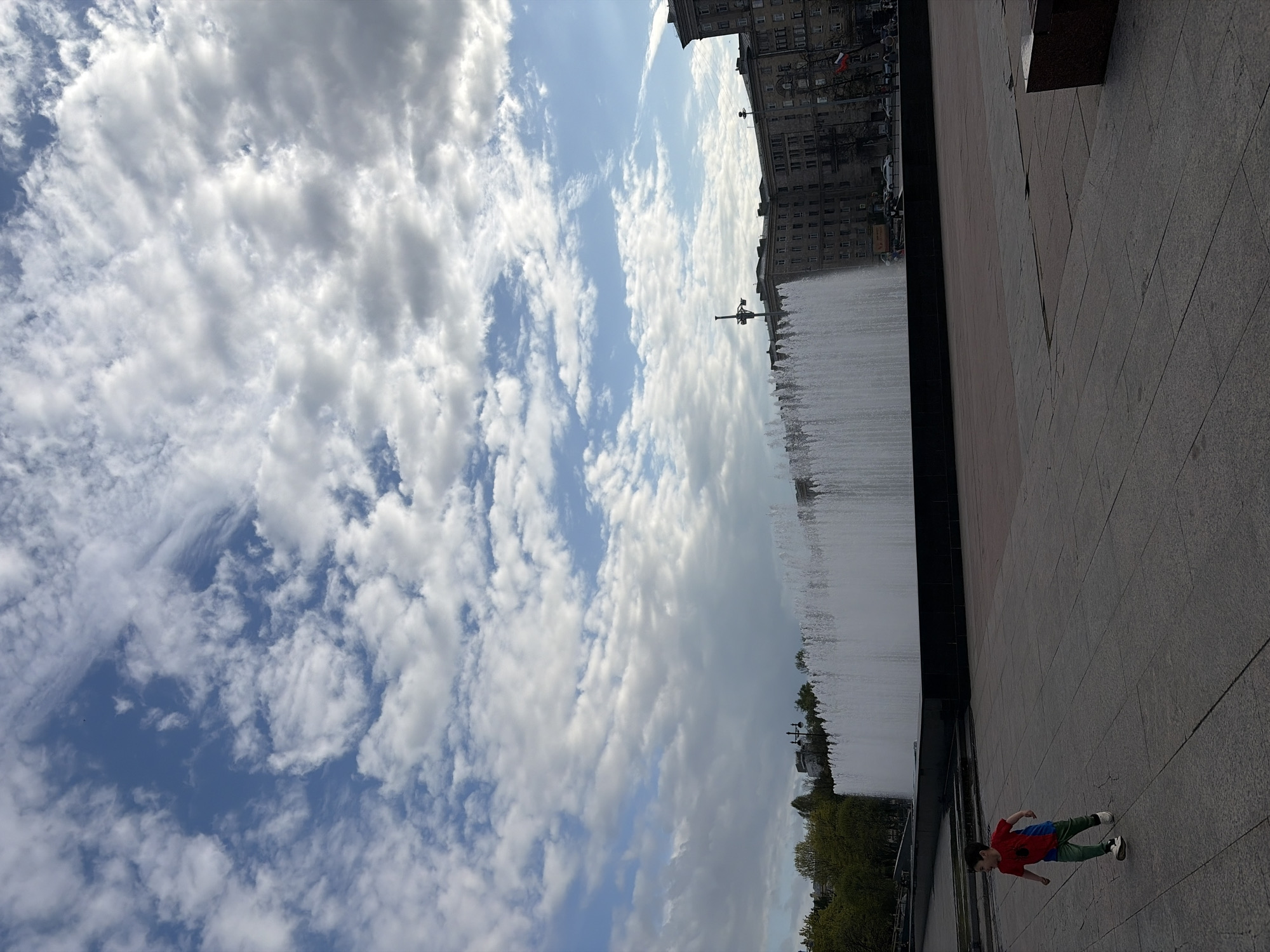 The image depicts a scene in Saint Petersburg, Russia, showing the cityscape, a large water fountain, and a child on a paved walkway. The sky is filled with numerous white, puffy clouds. Buildings are visible in the background near the fountain. The paved walkway is in the foreground, where a child wearing a red shirt and green pants is walking.