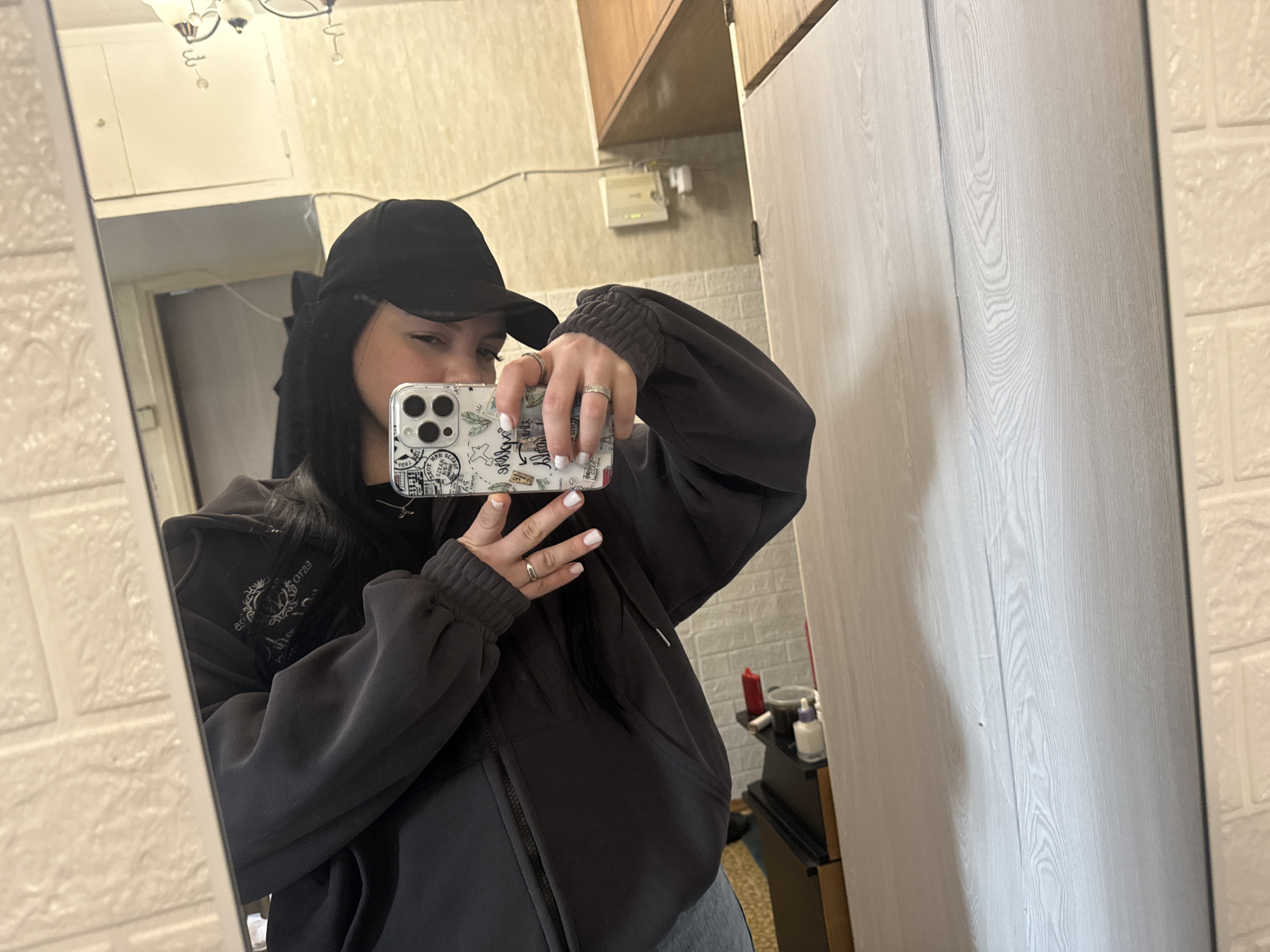 A woman in a black cap and a gray jacket is taking a selfie in a mirror. She is holding her phone up with her left hand. Her nails are painted white. A silver ring is on her ring finger and another one is on her index finger. The background shows a white wall with a brick pattern and what appears to be a cabinet and a shelf. The reflection in the mirror shows another room. The location cannot be determined by using the city of Mytishchi, Russia.