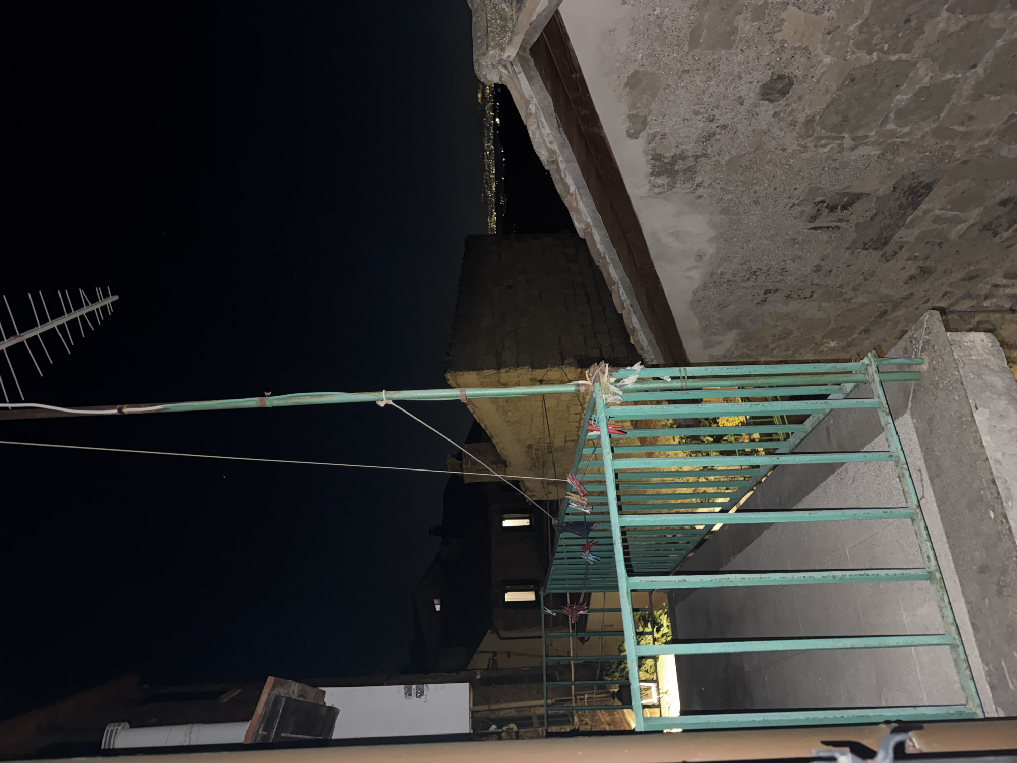 An outdoor scene taken at night in Castelnuovo di Porto, Italy. In the foreground is a metal railing painted light green. Clothes with clothespins are hung up to dry on the railing. A building overhangs the railing. In the background, buildings are dimly lit with windows visible. An antenna is seen against the night sky. Faint city lights are visible in the distance.
