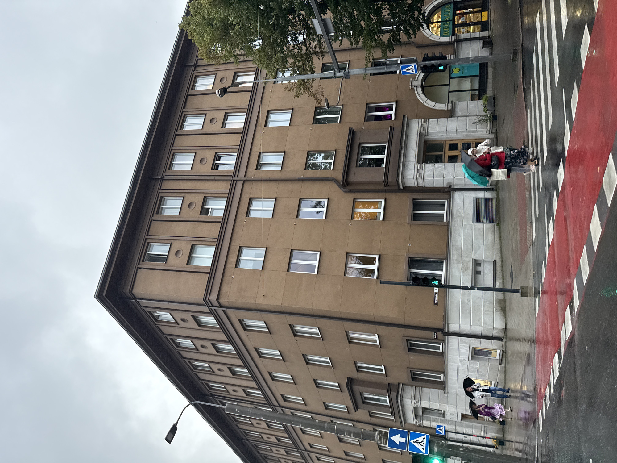 The image depicts a wide shot of a brown, multi-story building on a cloudy day in Tallinn, Estonia. The building has numerous windows and is situated on a street with a pedestrian crossing marked by red and white stripes. Several people are visible, crossing the street under umbrellas. One group of two individuals, one in a purple dress and the other in white, are walking under separate umbrellas. Another group of three individuals, two in the foreground and one behind, are crossing the street; one person is wearing a red skirt and holding a turquoise umbrella. Traffic lights are visible, and one appears to be green. A street lamp stands on the sidewalk. The overall environment suggests a rainy or damp day in an urban setting. The building's architecture suggests a mid-20th century style. A sign on the building indicates "REGALIA" above an entrance. The street signs read "Tõnismägi 1".