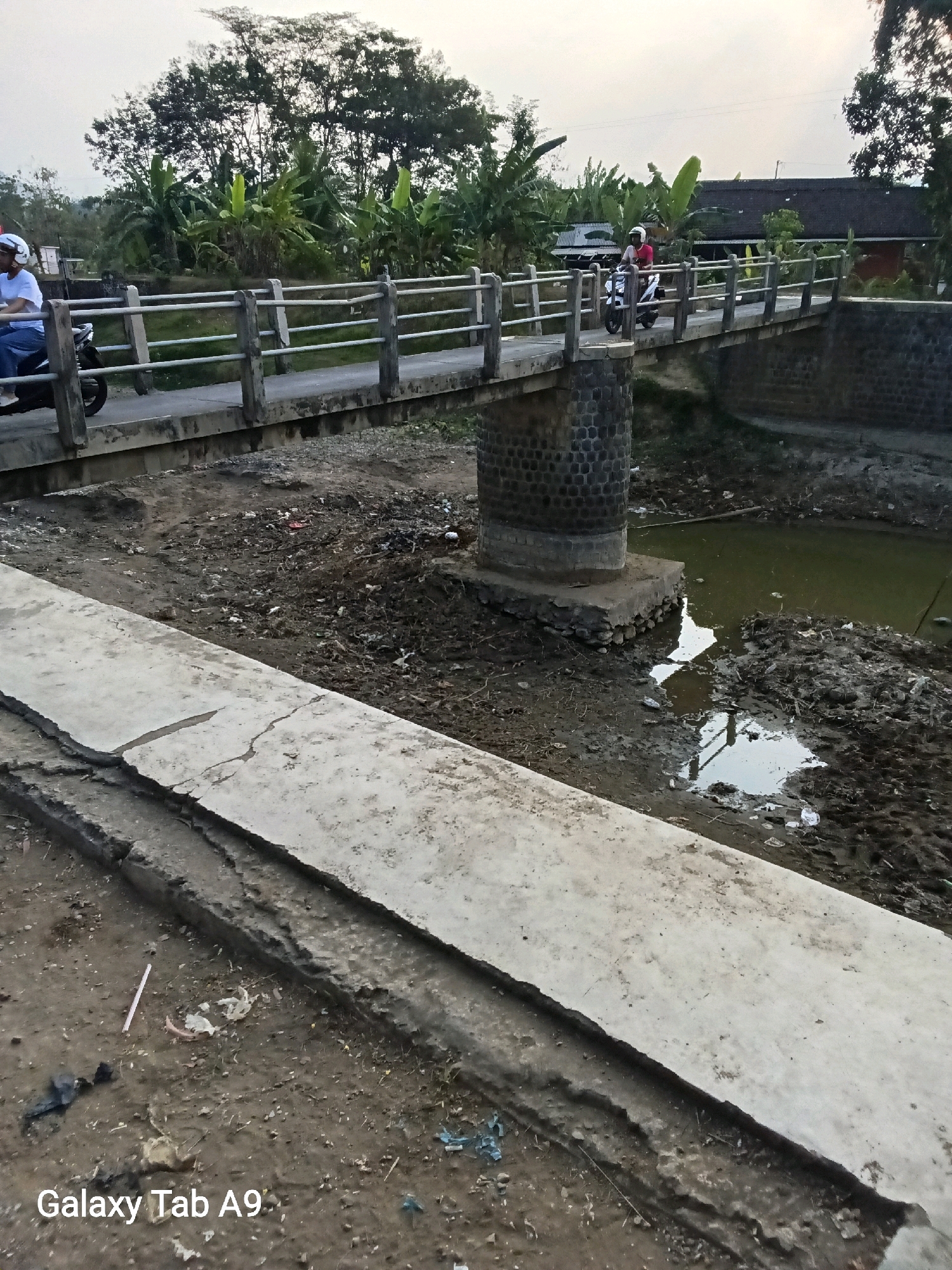 The image shows a bridge over a dry riverbed in Sumberingin, Indonesia. Two people are riding motorcycles across the bridge. The riverbed is filled with dirt and debris, and there is a small amount of water in one section. The bridge is made of concrete and stone, and it has a railing on either side. In the background, there are trees and houses. The sky is overcast.