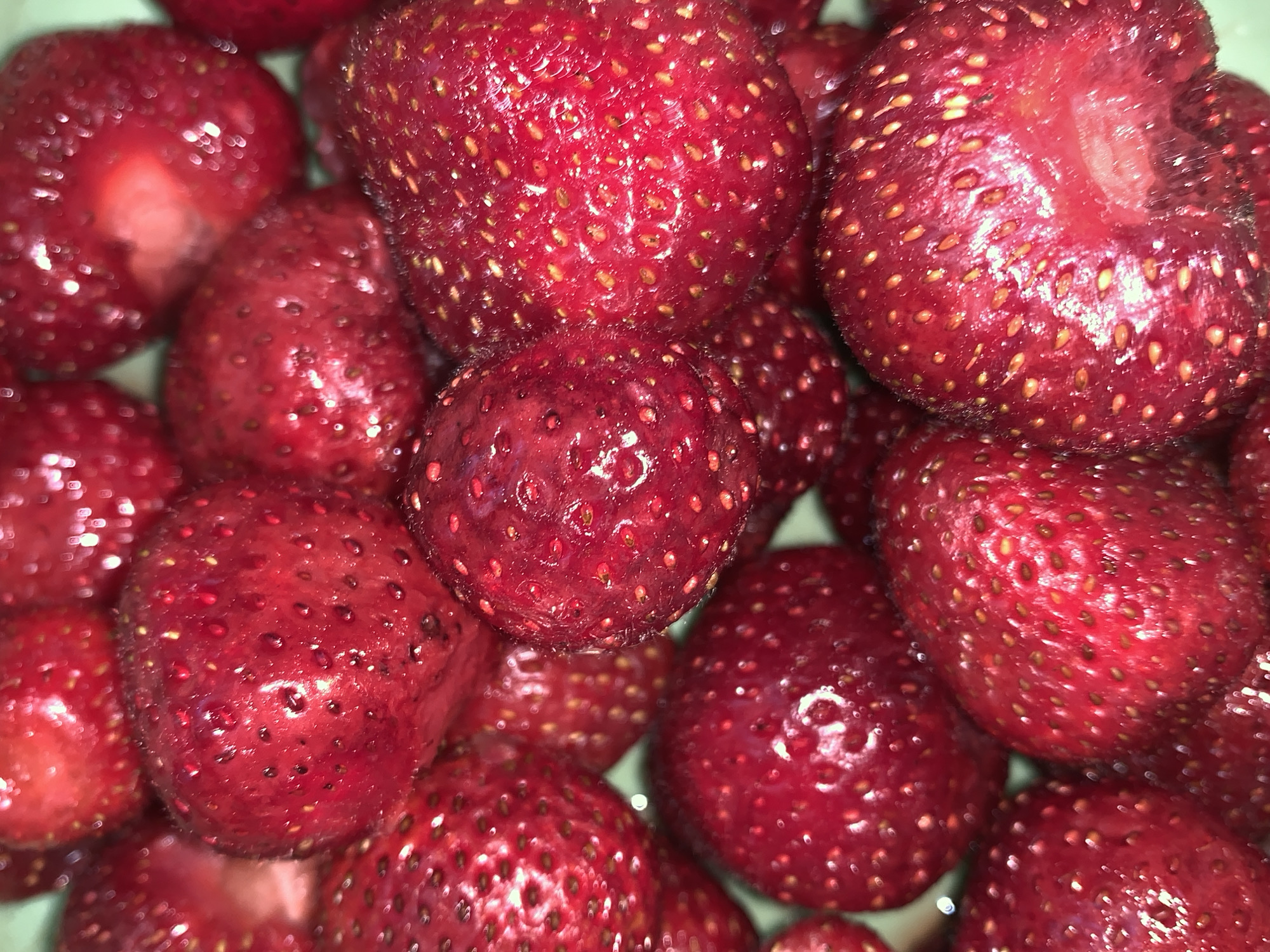 The image displays a close-up, overhead view of a pile of ripe strawberries. The strawberries are a vibrant red color, with numerous small, yellow seeds dotting their surfaces. Some of the strawberries appear to have been cut open, revealing their white flesh. The lighting suggests this is likely an indoor setting, possibly a kitchen or pantry, as there are reflections of light on the glossy surfaces of the fruit. The overall impression is one of fresh, abundant produce, likely prepared for consumption. There is no visible text in the image.
