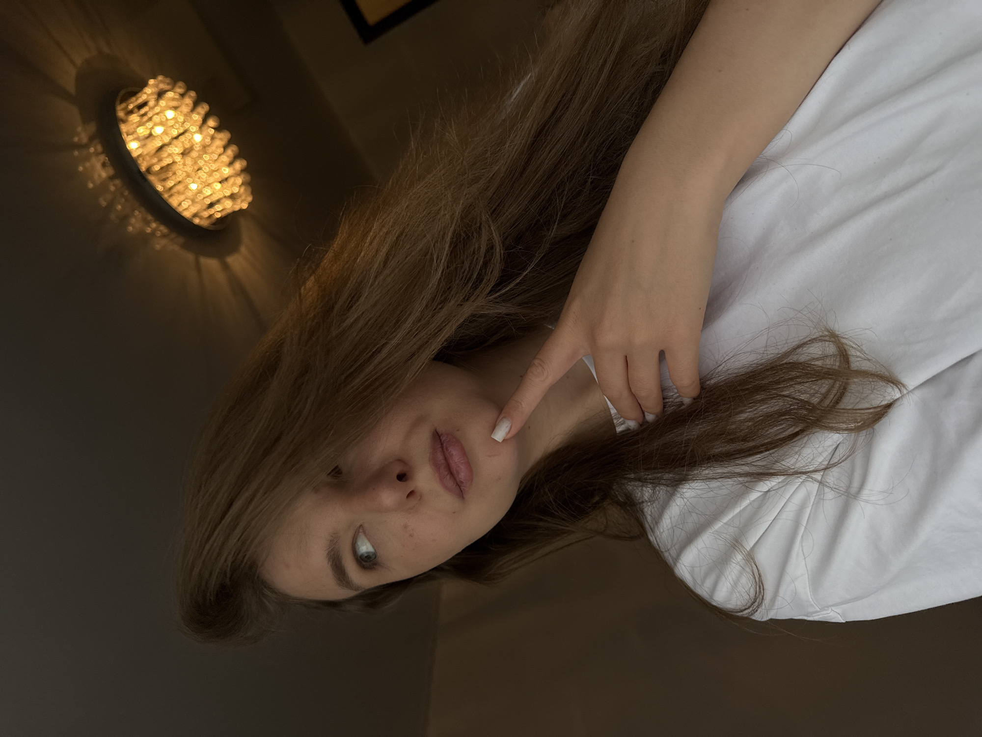 A young woman with long, light brown hair is captured in a close-up, slightly angled shot. She is wearing a plain white shirt. Her head is tilted, and her gaze is directed slightly away from the camera, giving her an introspective appearance. Her right hand is raised, with a single finger touching her cheek near her mouth. A ceiling light fixture with multiple small, bright bulbs is visible in the upper left corner of the frame, casting a warm glow and creating a pattern of light and shadow on the wall. The background appears to be an indoor setting with dark walls and a hint of a wooden frame on the wall above the light fixture. The lighting suggests it is likely evening or a dimly lit indoor space. The woman's expression is neutral, possibly thoughtful. There is no visible text in the image.