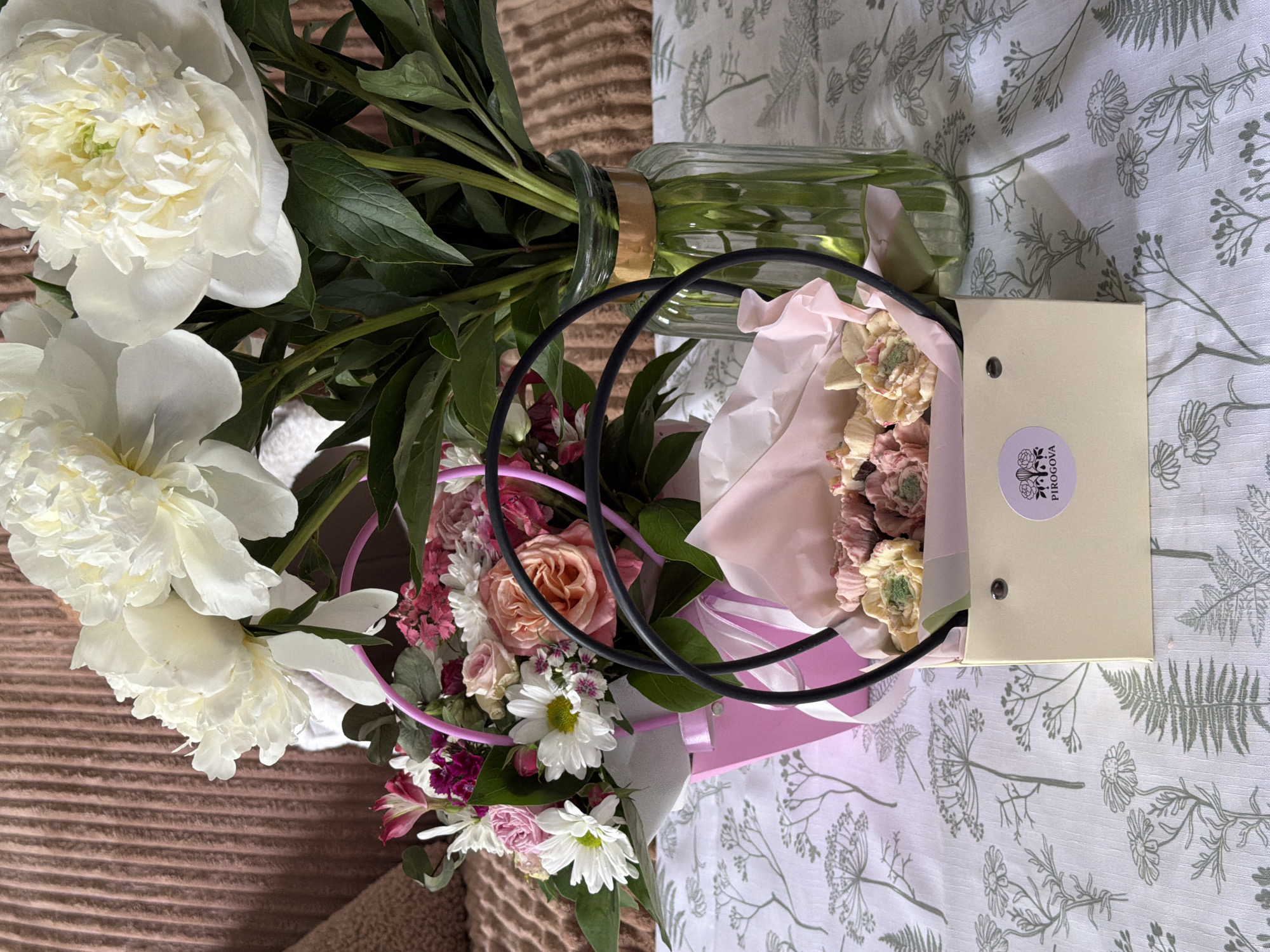 A bouquet of flowers is placed on a surface. The bouquet includes white peonies and a variety of smaller flowers in shades of pink, peach, and white. The smaller flowers are arranged in a light pink container with black handles. Some of the flowers are wrapped in white and pink tissue paper. Next to the bouquet is a tan rectangular box with a round sticker on it. The bouquet in glass vase lays on brown fabric next to the bouquet in the pink container. The background is a light fabric with a floral pattern. The exact location is not specified.