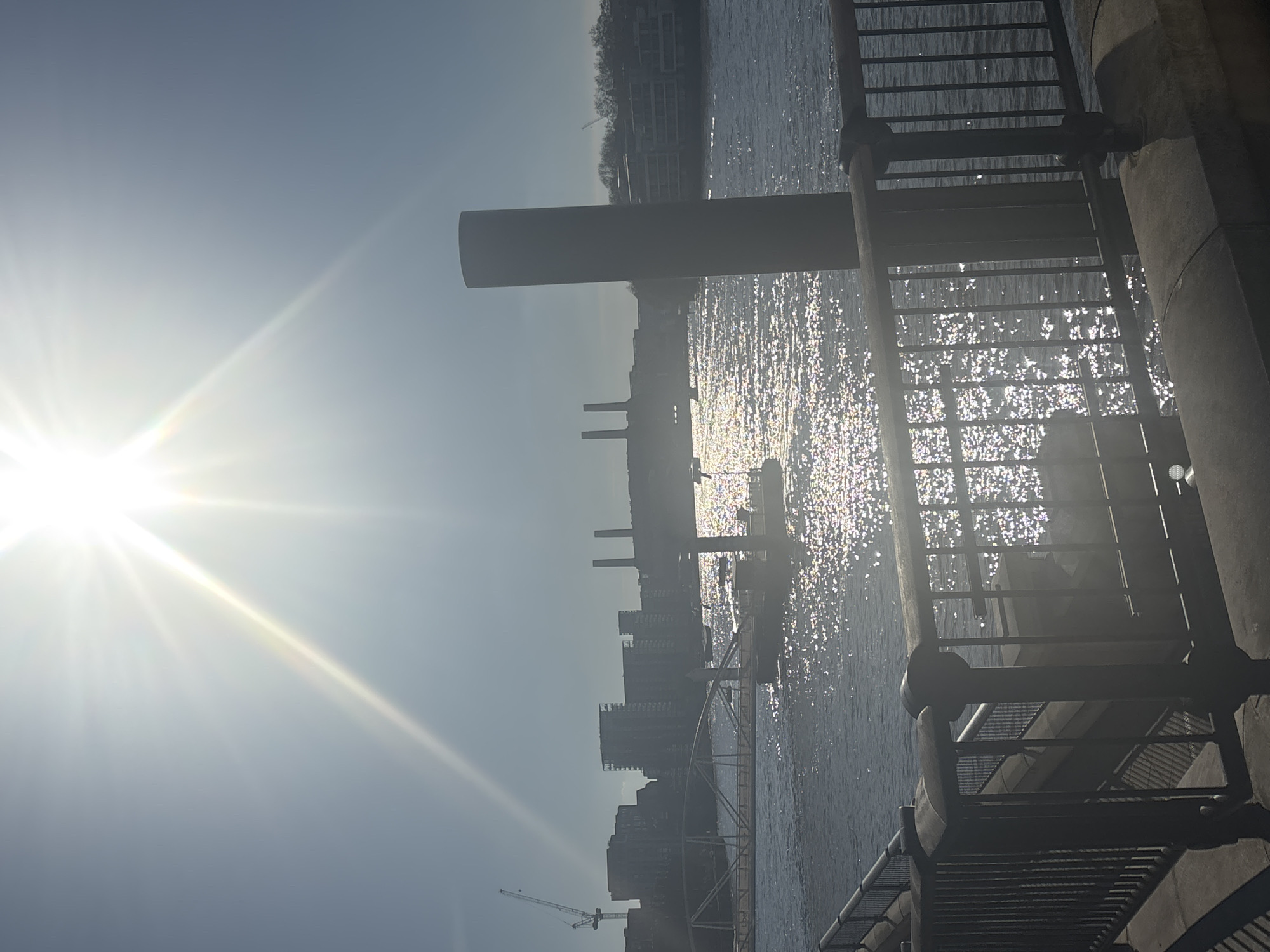 The image shows a bright, sunlit scene in London, United Kingdom, likely near the River Thames. The sun is positioned high in the sky, creating glare on the water's surface. Battersea Power Station can be seen in the background. A construction crane is visible in the skyline, suggesting ongoing building development. A metal structure, possibly part of a walkway or railing, occupies the foreground, casting shadows. The image is oriented sideways.
