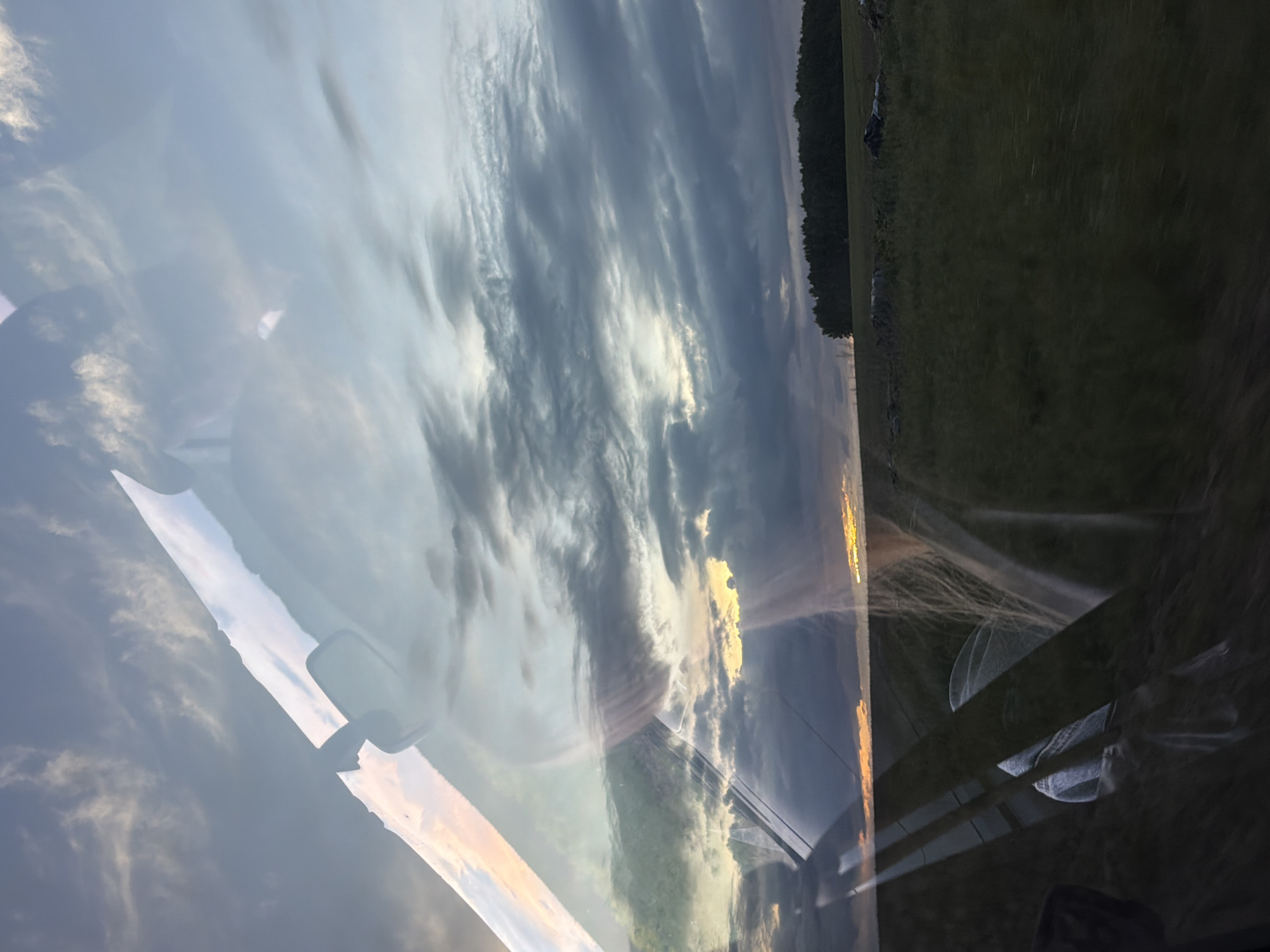 The image appears to be taken from inside a vehicle, looking out through the windshield. The sky is filled with clouds, some appearing dark gray while others are lighter, reflecting the sun. A strip of land is visible, featuring a line of trees. The reflection of elements from inside the vehicle can be seen on the windshield. The location is Kostovaty, Russia.