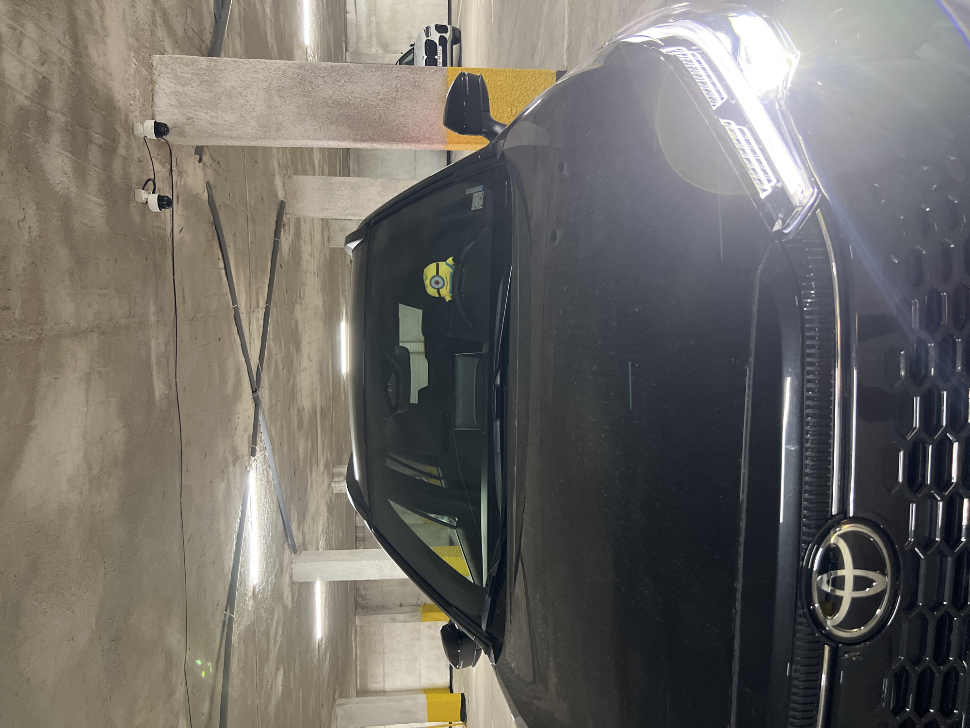 This is a close-up, frontal view of a black Toyota SUV parked in an underground parking garage. The car's headlights are on, casting a bright glare. A stuffed Minion toy is visible on the dashboard, sitting on the driver's side. In the background, a white BMW SUV is parked further down the aisle. The parking garage has a concrete ceiling with exposed pipes and fluorescent lighting, and reinforced concrete pillars with yellow markings. The scene appears to be in the city of Vratsa, Bulgaria.