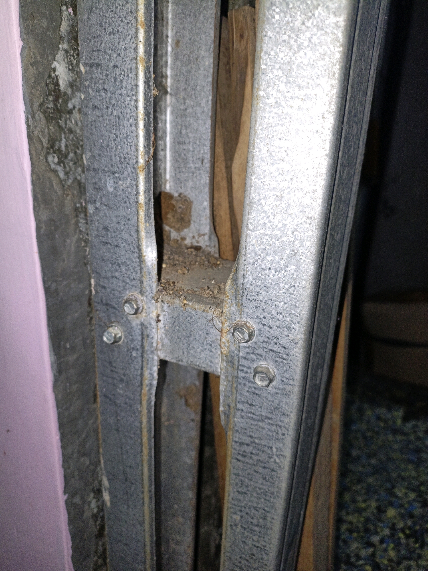 A close-up, ground-level shot shows a section of a metal door frame, likely a garage or utility entrance, situated in Cikal, Indonesia. The frame is constructed from corrugated galvanized steel and exhibits signs of rust and wear, particularly around the bolt connections. The left side of the frame is adjacent to a wall covered in a pale pink material, which appears to be some form of insulation or cladding. The background reveals a wooden surface, possibly part of the door or an underlying structure, and a dark, speckled floor. The lighting is dim, suggesting an indoor or shaded outdoor environment. The scene depicts a common urban or suburban architectural element, highlighting material condition and environmental exposure.
