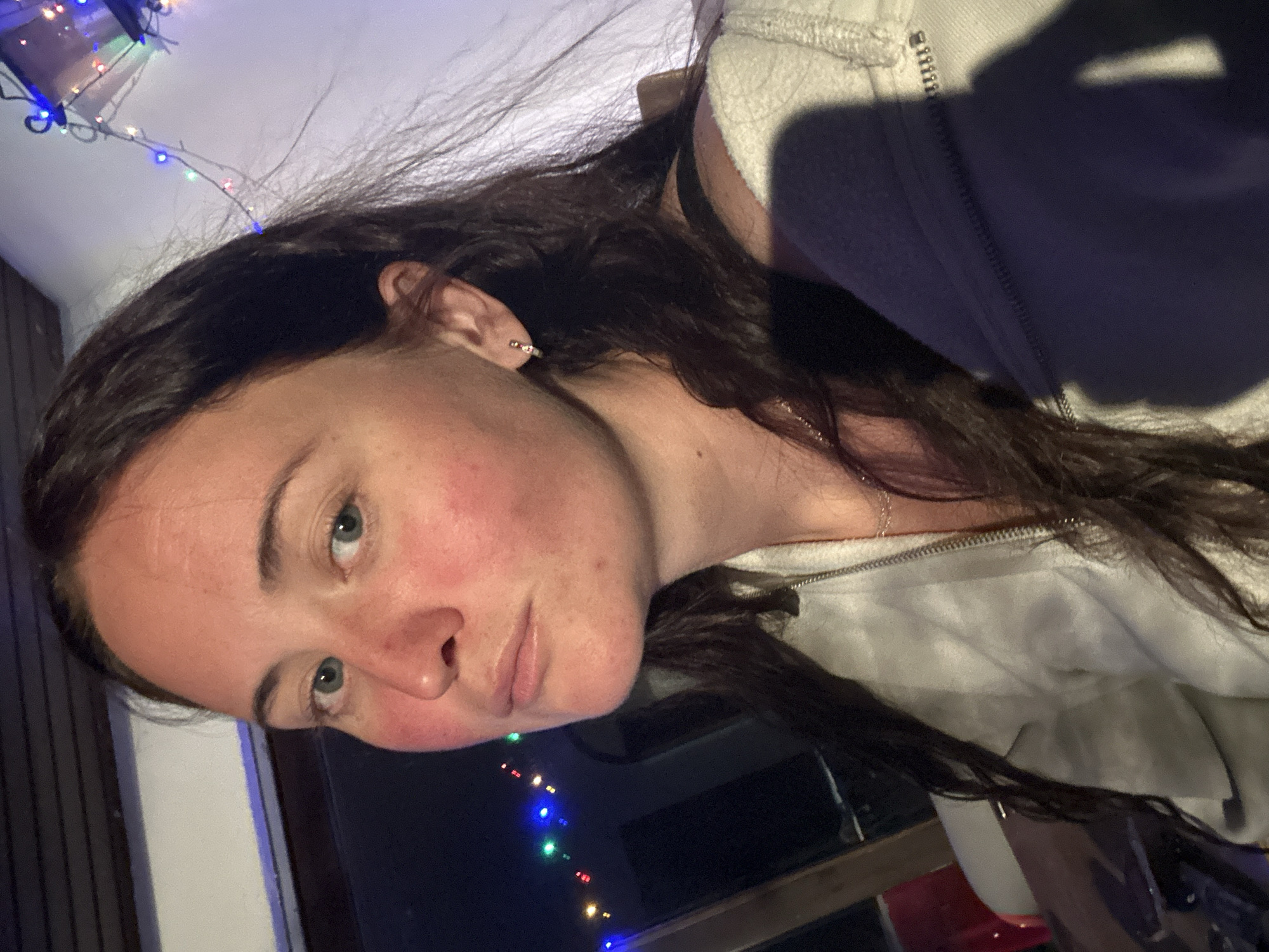 A young woman with long, dark brown hair is pictured from the chest up, looking towards the camera. She has fair skin that appears flushed on her cheeks and nose, and her eyes are a greenish-blue color. She is wearing a light-colored zippered hoodie, and a delicate necklace. A small hoop earring is visible in her right ear. Behind her, colorful string lights are strung across a dark background, suggesting an indoor setting possibly decorated for a holiday or celebration. The lighting in the scene is dim, with some areas illuminated by the string lights and a visible shadow cast on her hoodie.