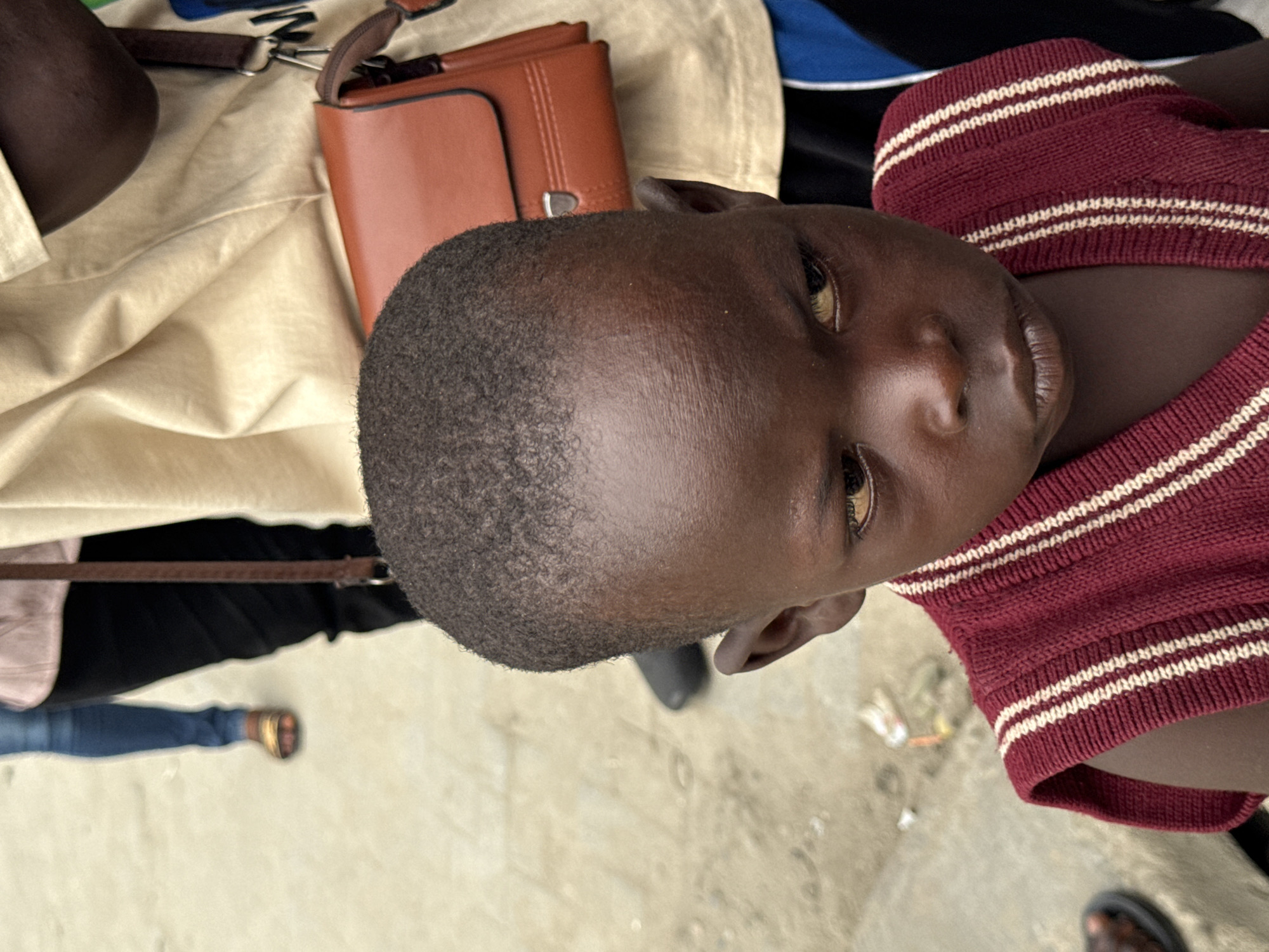 A child with short hair is seen from an overhead perspective, wearing a burgundy and white striped shirt. They are surrounded by objects including a tan purse, a brown leather bag, and a black shoe. In the background, partial views of black pants, blue jeans with brown sandals, and a black sandal are visible on a concrete or paved surface. The location is unspecified; there is no explicit indication of Lagos, Nigeria.
