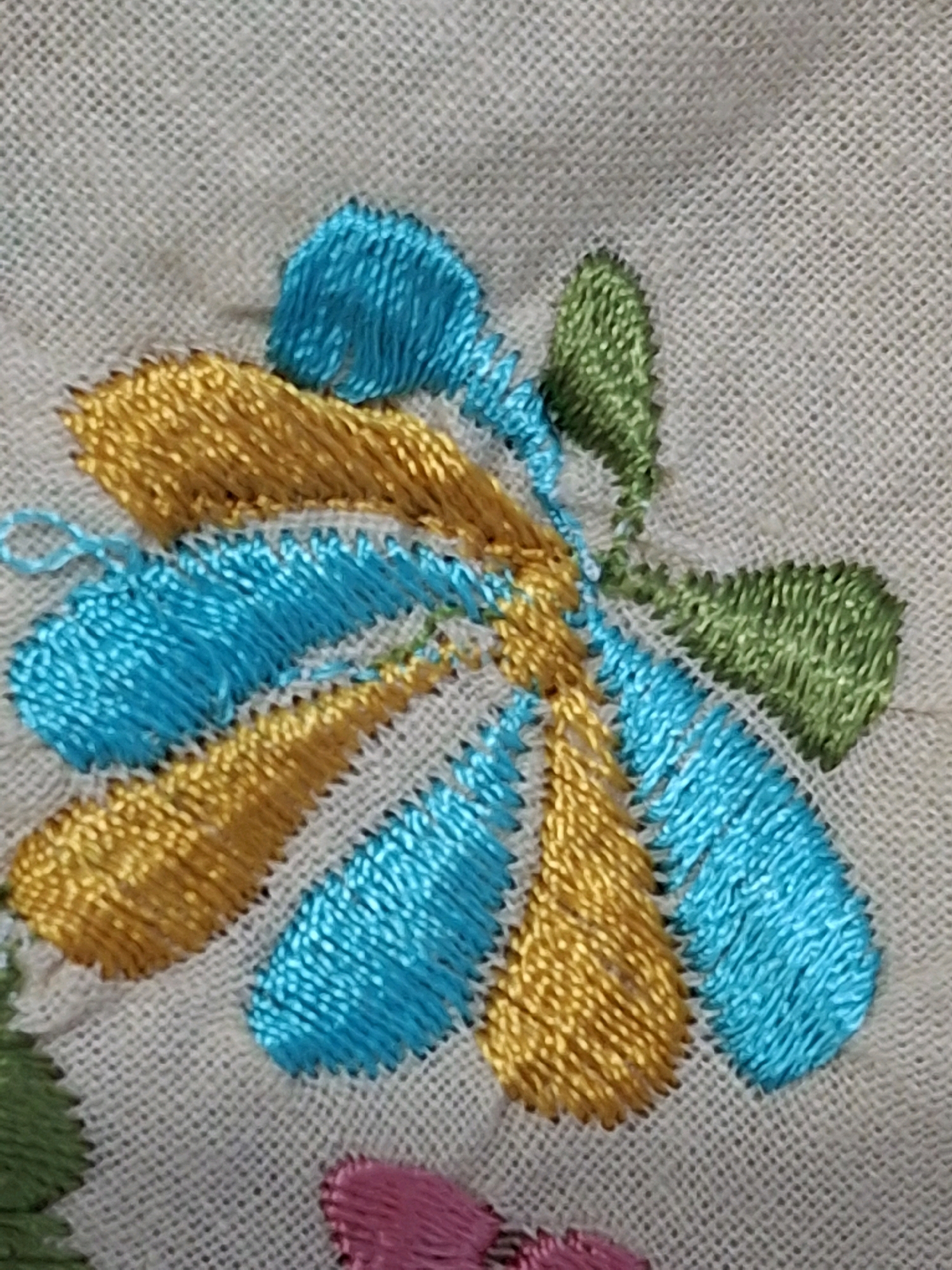 A close-up, overhead shot shows a portion of embroidered fabric. The embroidery depicts a stylized flower with several teardrop-shaped petals in alternating shades of teal and gold, interspersed with green petals. The fabric itself appears to be a light-colored, woven material. The image is a still photograph and there are no people, actions, or interactions to describe. The location of this image in Lumajang, Indonesia, is not discernible from the visual information provided.