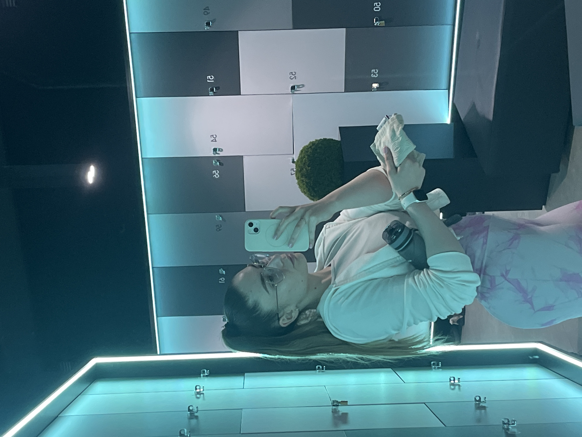 The image shows a woman with long brown hair in a ponytail taking a selfie in what appears to be a locker room. She is wearing glasses, a white long-sleeved shirt, and pink and white leggings. She is holding a white smartphone in her left hand and a water bottle in her right hand. Behind her is a wall of lockers with numbers on them, arranged in a checkerboard pattern of gray and white. There is a small green bush on top of one of the lockers. Below her, there appears to be a shiny, turquoise-colored surface with visible locks on the lockers. The location is in an unknown city.