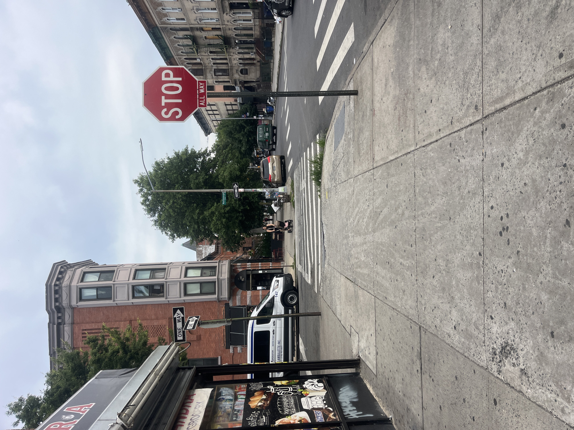 This is a street-level, vertical photograph taken from a sidewalk in New York City. The image is oriented such that the sky is on the left and the ground is on the right. A red octagonal stop sign with "STOP ALL WAY" in white lettering is prominently displayed near the center of the frame. Adjacent to it is a street sign indicating "McCullough".
The scene depicts an urban intersection with a crosswalk marked by white stripes. Several vehicles are visible, including a white van with "OPERATOR DOT# 31522 AMBULETTE INC." printed on its side, a light-colored car, and a green vehicle further down the street. In the background, multi-story brick buildings with numerous windows line the street. Trees provide greenery in the middle ground, and people can be seen on the sidewalk near the intersection, some possibly waiting to cross. A street lamp is also visible.
A storefront with signage is partially visible in the foreground, featuring images of food and text, including "GROCERY" and "BREAKFAST". The concrete sidewalk appears worn and cracked in places. The sky is overcast, suggesting a cloudy day. The overall impression is a typical urban street scene in New York City, likely during daylight hours.