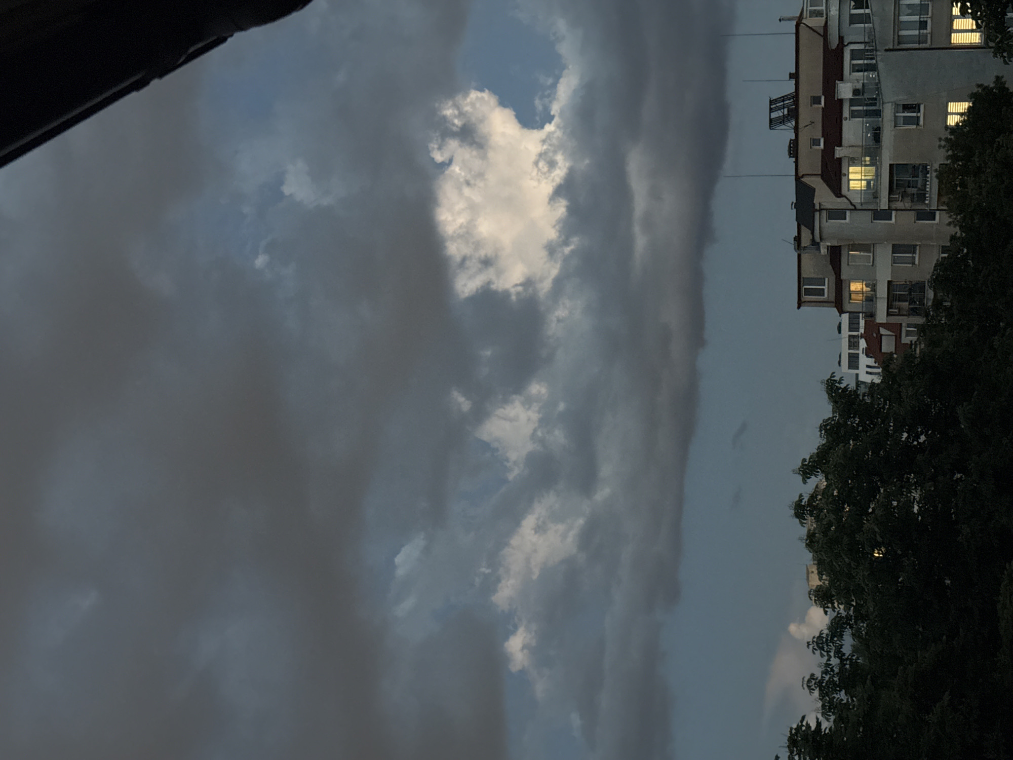 The image shows a cloudy sky over Sofia, Bulgaria. The sky is predominantly gray, with a patch of brighter, white clouds visible in the center. A building with lit windows is visible to the right, partially obscured by trees. Part of a roof can be seen in the upper-left corner. The overall scene appears to be taken during dusk or twilight.
