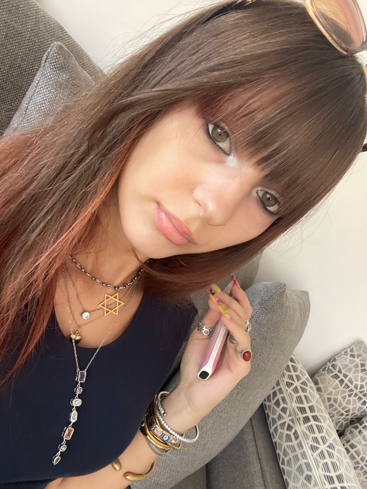 A young woman with long, brown hair and bangs is shown from the chest up, looking at the camera. She is wearing a dark blue top and multiple necklaces, including a Star of David pendant and a long pendant with colorful stones. Her fingernails are painted with bright colors. She is holding a pink and white electronic cigarette in her right hand, and wearing several rings on her fingers. She is sitting on a gray couch, with pillows on either side. The lighting suggests an indoor setting, possibly daytime. The image does not provide any specific clues about the location within Tel-Aviv, Israel.