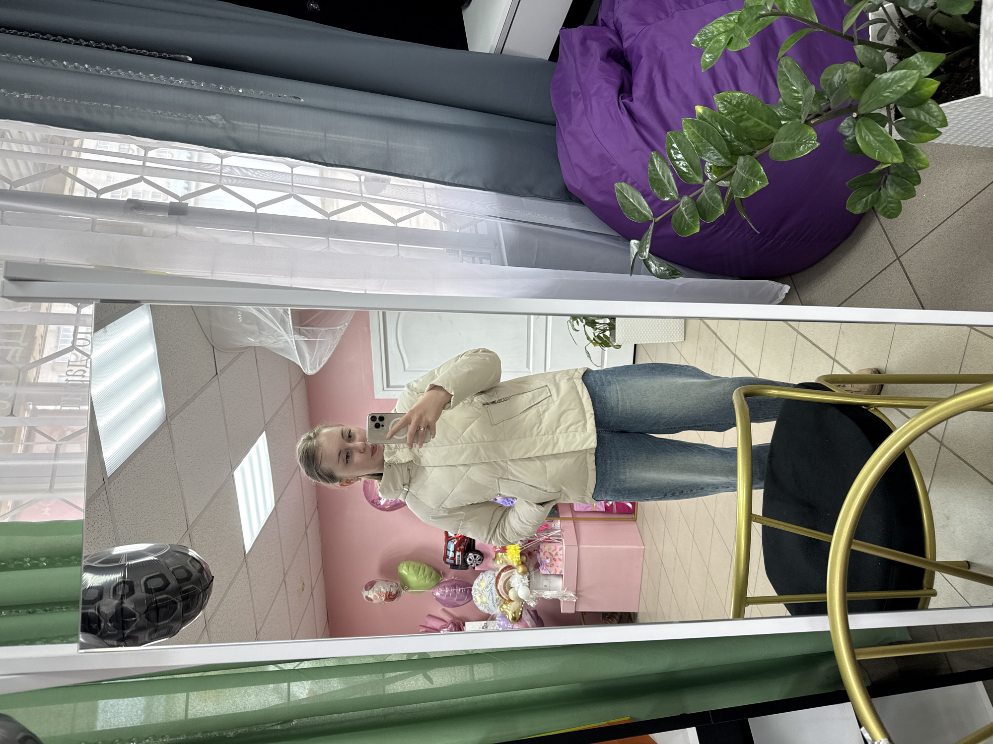 The image shows a reflection in a mirror of a person holding a cell phone, presumably taking the picture. The person is wearing a light-colored puffer jacket and blue jeans. Behind the person in the reflection is a pink wall adorned with balloons, and a pink table with what appears to be a gift display. In the real space, outside of the mirror’s reflection, there is a purple beanbag chair, a green plant, and a chair with a black seat and gold frame. There are also blinds of different colors (grey, white, and green). The exact location in the city of Unknown, Unknown cannot be determined from the image.