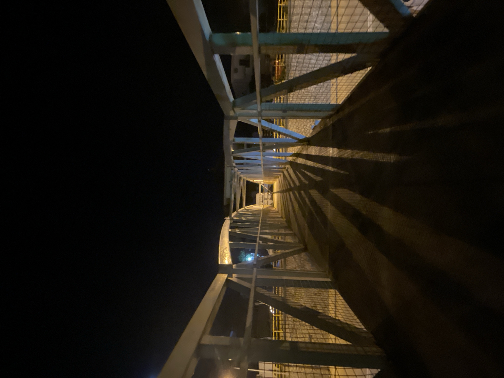 The image shows a pedestrian bridge at night in the city of Kassandra Municipal Unit, Greece. The bridge is made of white metal with diagonal supports and a wire mesh fence on the sides. Long, dark shadows are cast across the bridge's walkway from the bridge's structure, indicating a light source behind the photographer. The sky is dark, and the surrounding environment is dimly lit by artificial lights, with some buildings and yellow railings visible in the background. There are no people or animals visible in the image.