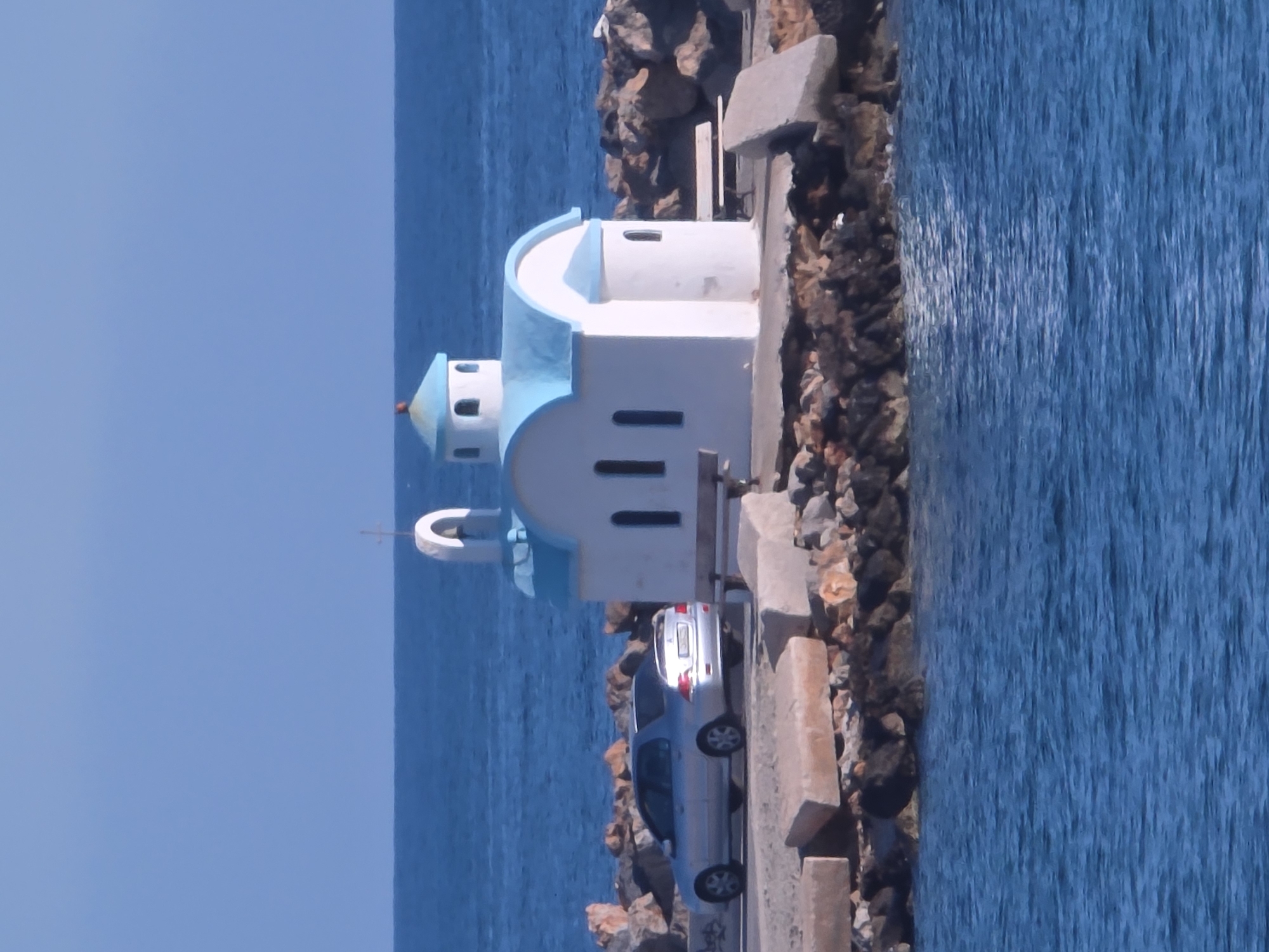 A small, white chapel with a bright blue dome and roof sits on a rocky breakwater in Chania, Greece. The chapel has a bell tower with a cross on top and arched windows. A silver sedan is parked to the left of the chapel. Benches are visible on either side of the chapel. The sea, a deep blue, surrounds the breakwater. The sky is clear and blue.