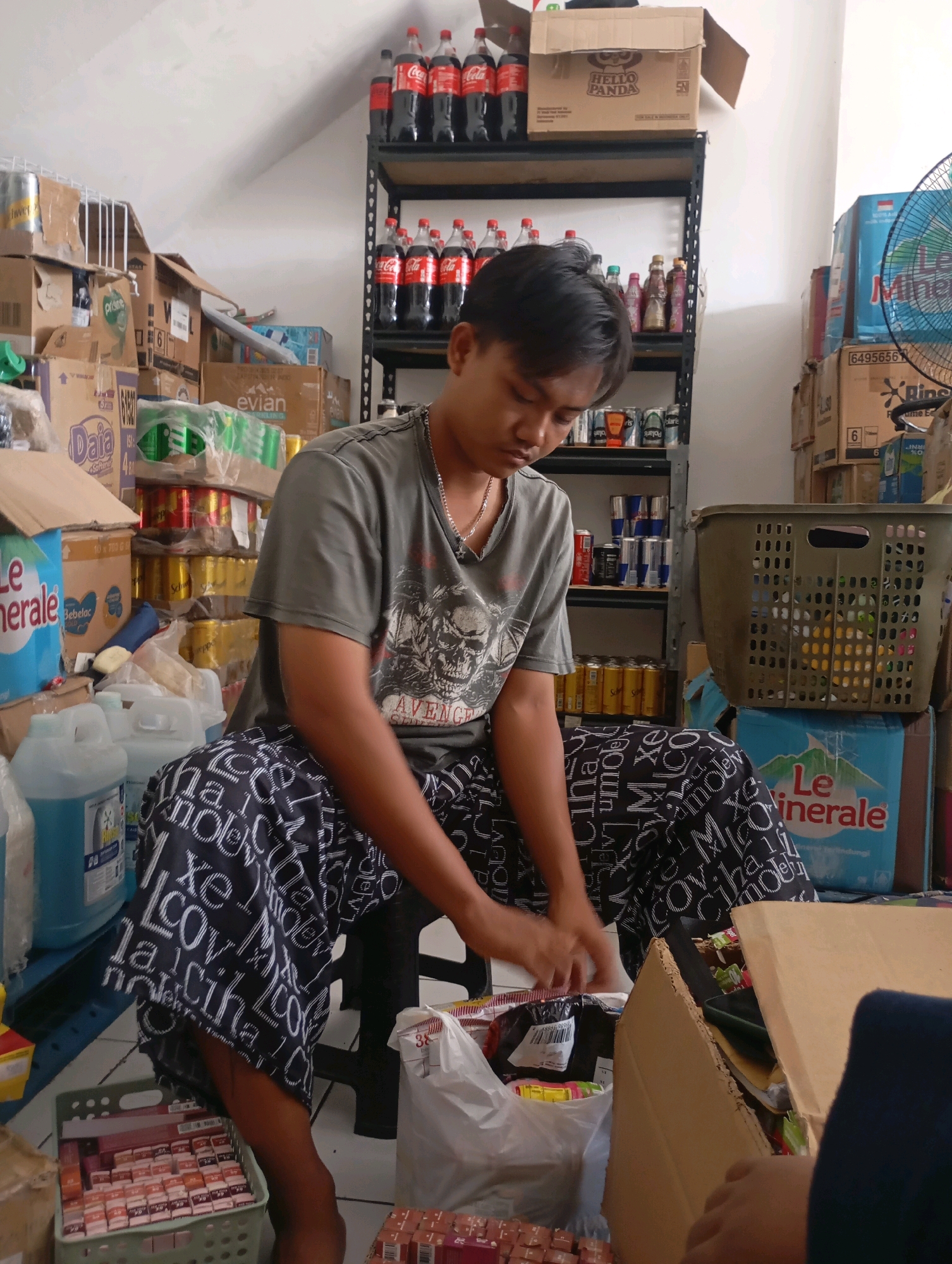 A young man in a grey t-shirt with a skull print and patterned pants is sitting in a store filled with goods. He is looking down and appears to be handling some items, possibly organizing or packing them. The store is packed with shelves full of drinks in bottles and cans, as well as boxes and other assorted merchandise. A fan is visible in the background. The scene suggests a busy retail environment, likely a small grocery or convenience store. The presence of goods like Coca-Cola and Le Minerale suggests this store could be located in an area where these brands are commonly sold, such as Babelan, Indonesia.