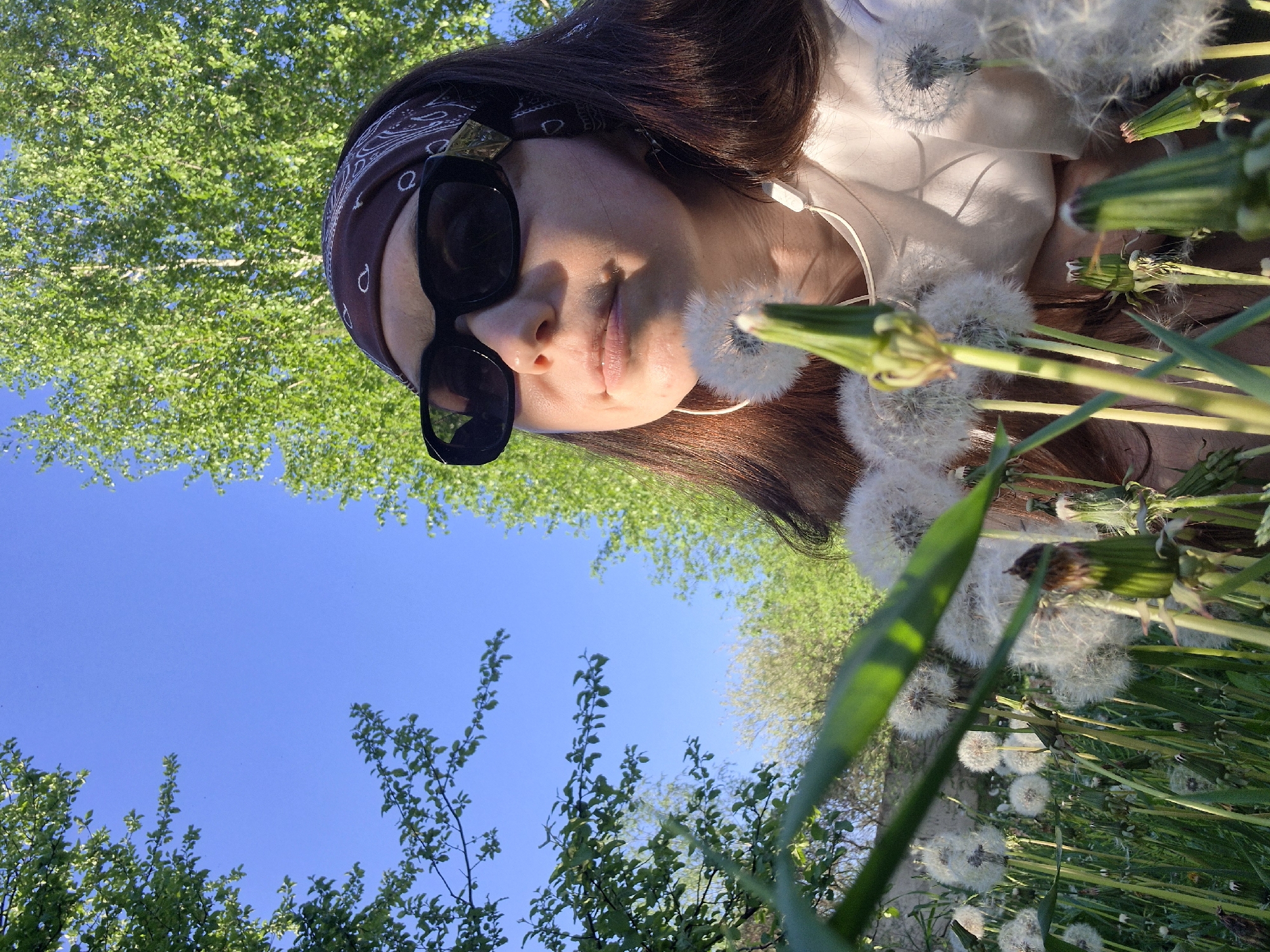 The image shows a person lying in a field of dandelions. The person is wearing sunglasses and a bandana, and has headphones in their ears. The sky is blue and there are trees in the background. The location is Unknown, Unknown.