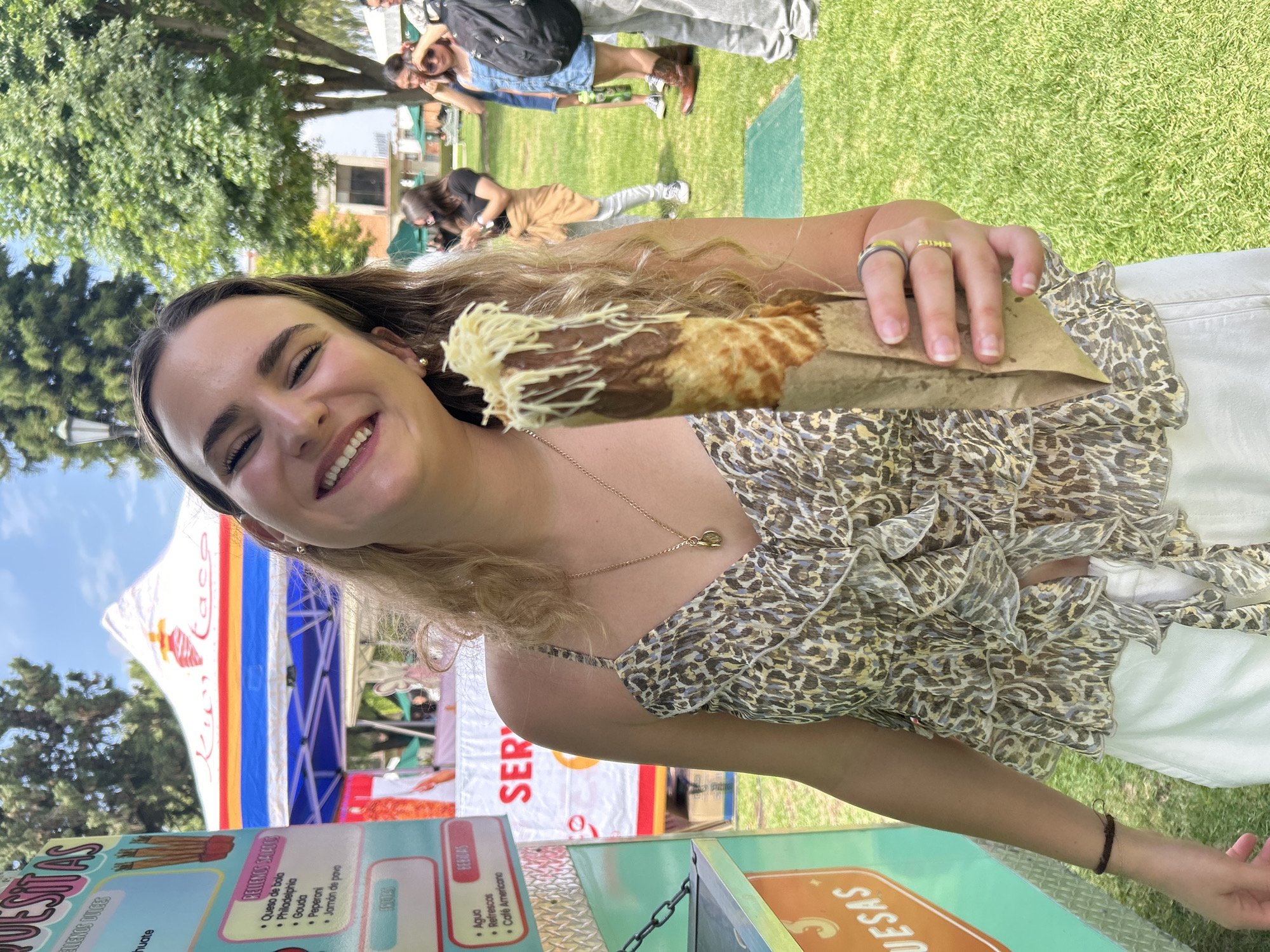 The image shows a young woman smiling while holding a food item, likely a crepe or quesadilla with cheese, wrapped in paper. She is outdoors, as suggested by the presence of a grassy area and trees in the background. Behind her is a food stand or cart named "Quesitas" with menu items listed, and a canopy with red, white, and blue stripes. Other people can be seen in the background, suggesting a public park or fair-like environment. The food stand seems to offer items like quesadillas with various fillings (cheese, Philadelphia, Gouda, pepperoni, ham) and beverages. The location appears to be in the city of Unknown, Unknown.
