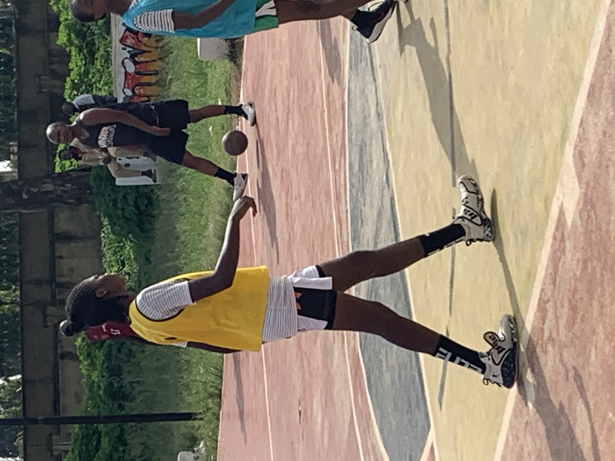 The image shows a group of people on an outdoor basketball court. The main subject is a young woman wearing a yellow vest over a striped shirt and black shorts, with black and white basketball shoes. She is positioned in the foreground, facing left. In the background, several other individuals are visible, some wearing athletic attire. One man in a black tank top and shorts is seen in the middle ground, and another person in a blue shirt is in the upper left portion of the frame. The court has distinct color sections of red, yellow, and blue/gray, suggesting an urban or recreational setting. The presence of shadows indicates it is daytime, likely with sunny weather. The scene appears to be a casual basketball game or practice session, possibly in a park or community sports area. There is some graffiti visible on a wall in the background, with the letters "AFRICA" and "KING" seemingly present. The overall mood appears energetic and active, consistent with a sports event. The location is identified as Gura Top, Nigeria.