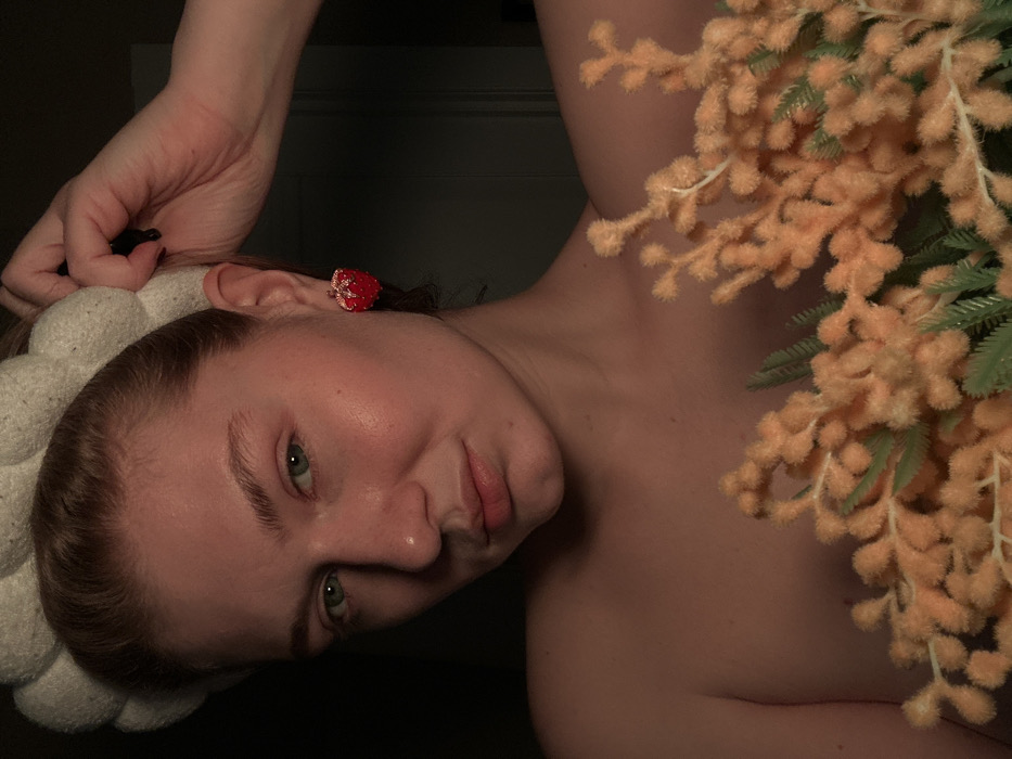A woman with a white, puffy headband tilts her head, revealing a red strawberry-shaped earring. Her face is fair, and her eyes are green. Her hand is raised, holding a black object near her head. Her shoulder is bare, and a cluster of fluffy orange flowers with green stems sits near her body. The lighting is low, casting a soft glow on her skin.