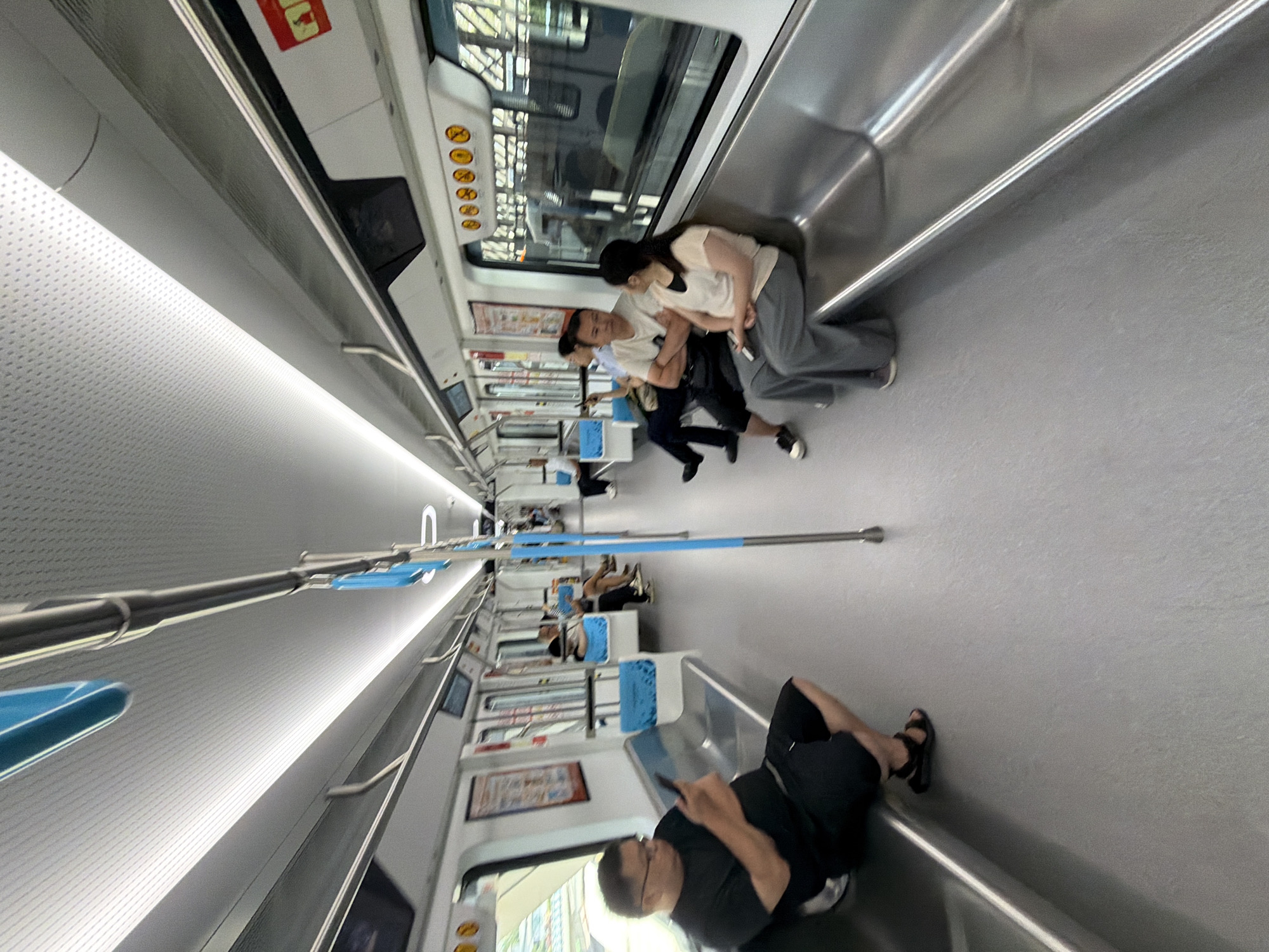 The image depicts the interior of a modern subway train, likely in Jiangjin, China. The train cars are spacious and brightly lit with fluorescent lighting along the ceiling. The seats are made of a silver-colored metal, with some sections featuring blue cushioned seating. Several passengers are visible, all seated.
In the foreground on the left, a man wearing a black t-shirt and black shorts is looking down at his phone. He is wearing black sandals. Further down the car on the left, a person in a white shirt and blue shorts is seated.
In the right car, a man in a white t-shirt and dark pants is seated with his arms crossed. Next to him, a woman in a light-colored top and grey pants is seated. Another person is seated further down the car, partially obscured.
The environment appears clean and well-maintained. There are advertisements and signage on the walls of the train cars, including a red and yellow warning sign on the right. The windows of the train show a blurred view of the outside environment, suggesting the train is in motion. The overall impression is one of a typical urban commute on public transportation.