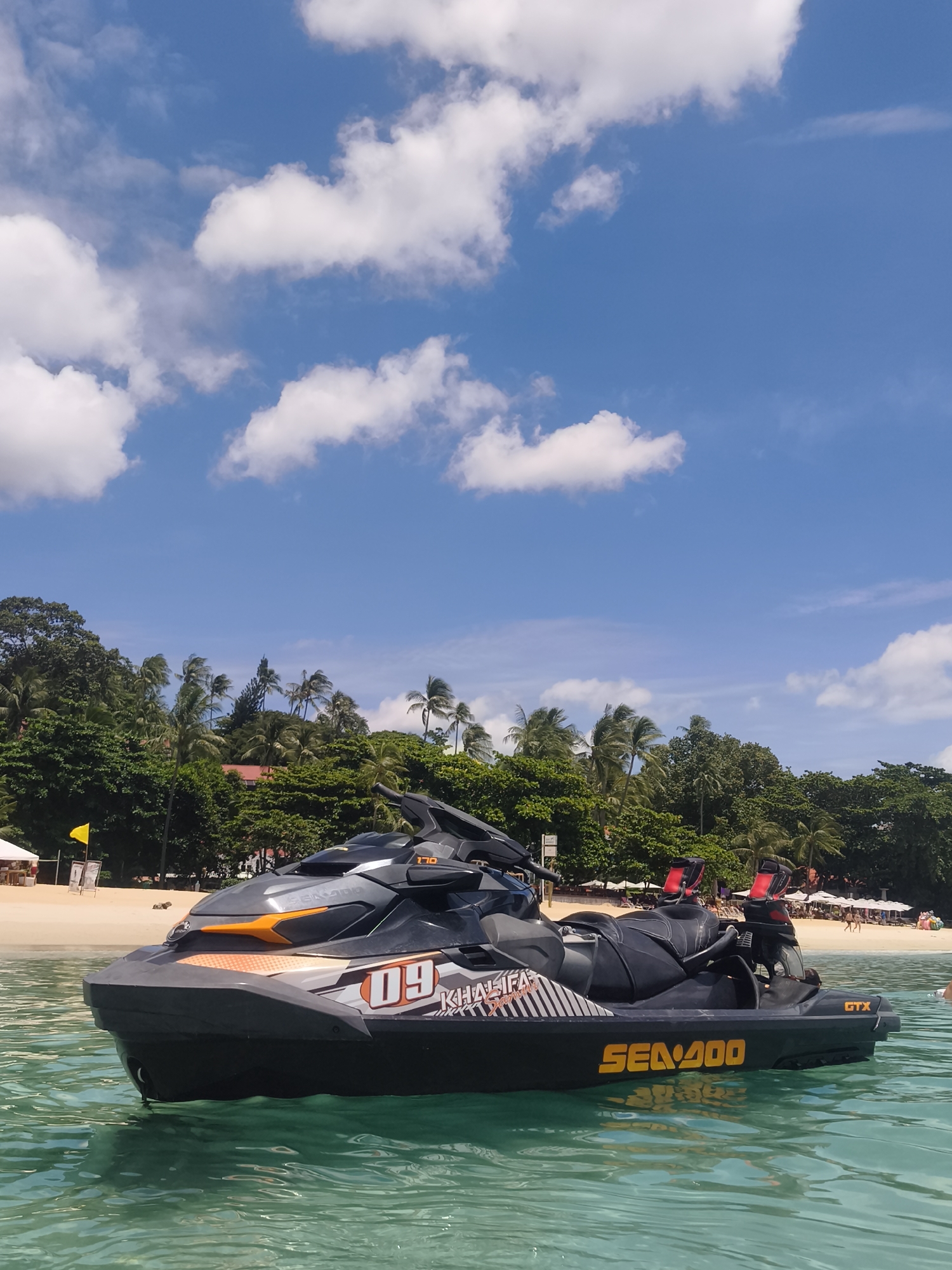 A black and orange jet ski is floating on turquoise water in front of a beach lined with palm trees and umbrellas. The sky above is blue with scattered white clouds. The jet ski has the number "09" and the word "KHALIFA" printed on its side. In the background, people are visible on the beach. The scene suggests a tropical vacation setting, possibly on the coast of Ko Samui District, Thailand.