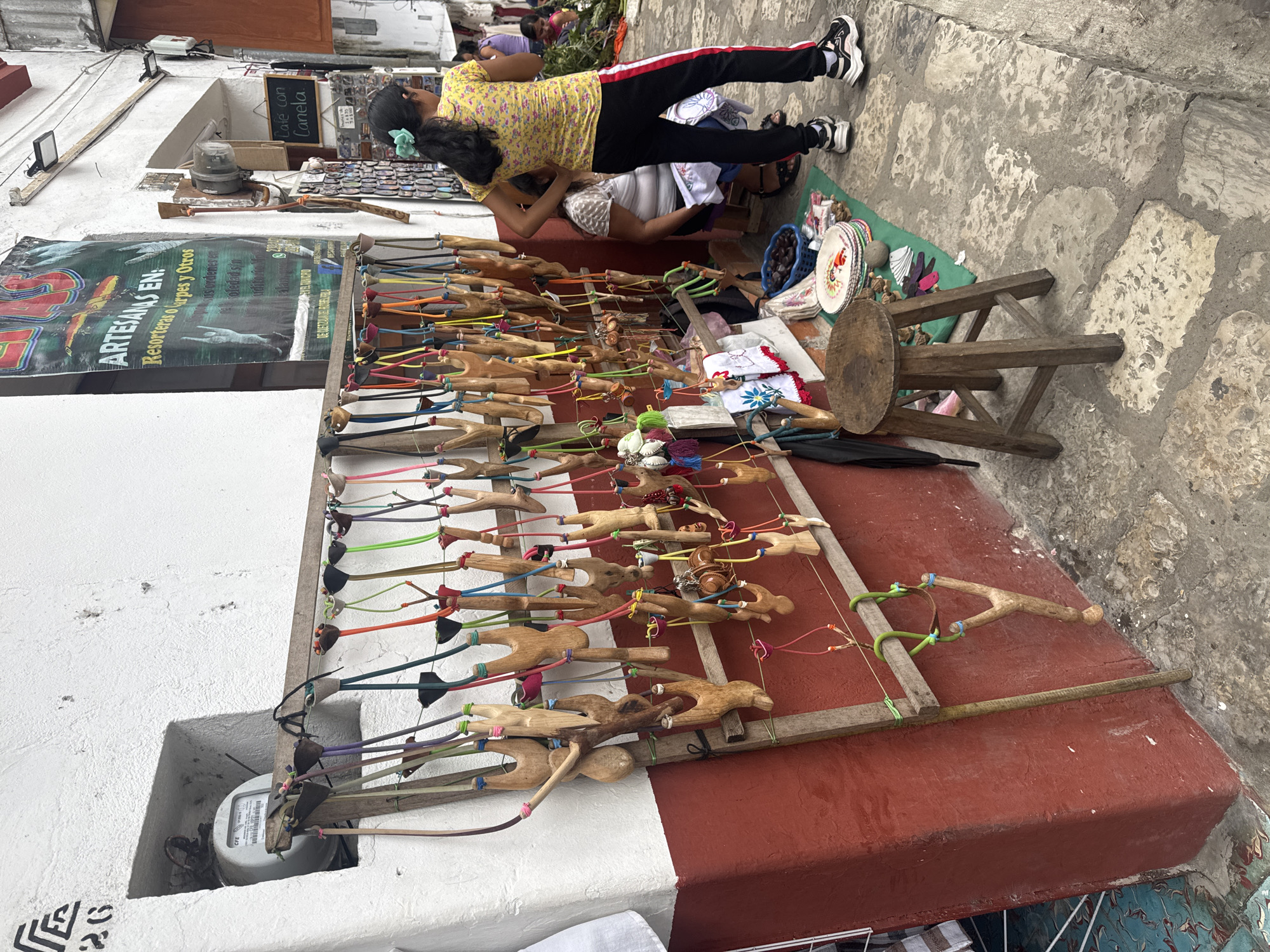 The image depicts a vendor's display of handcrafted wooden slingshots outside a shop in the city of Cuetzalan, Mexico. The slingshots, arranged in rows, feature various designs and colored rubber bands. Two people stand near the display; one is bending down as the other assists them. A wooden stool sits beside the display. A poster is affixed to the wall above the slingshots, and there are signs. The scene takes place on a stone-paved sidewalk next to a building painted white and red.