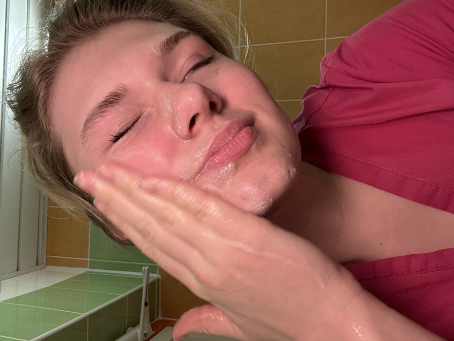 A fair-skinned person with closed eyes is applying a white, creamy substance to their face with their right hand. The person wears a pink top. The background features beige and green tiled walls and a glimpse of a window. The hand covers part of the cheek and appears shiny, suggesting moisture or the applied product. The individual's expression is serene.