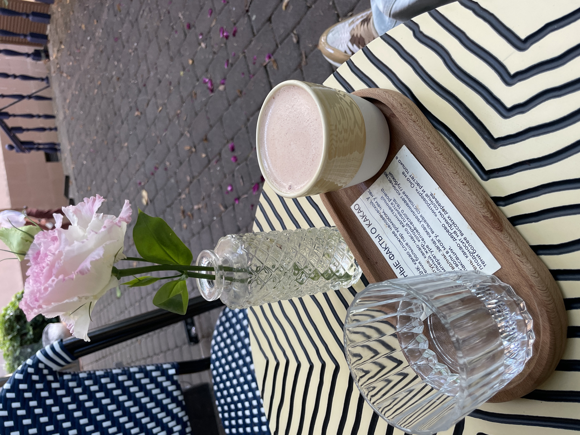 An outdoor cafe scene in Sochi, Russia, features a small table with a decorative zigzag pattern. On the table, a clear glass vase holds a single pink and white Eustoma flower. Next to it is a glass of water and a wooden tray holding a light yellow mug filled with a frothy beverage, likely hot chocolate, and a small piece of paper with text. In the background, a person's legs are visible, wearing casual sneakers. The ground is paved with cobblestones, and fallen petals are scattered around, suggesting a park or garden setting.