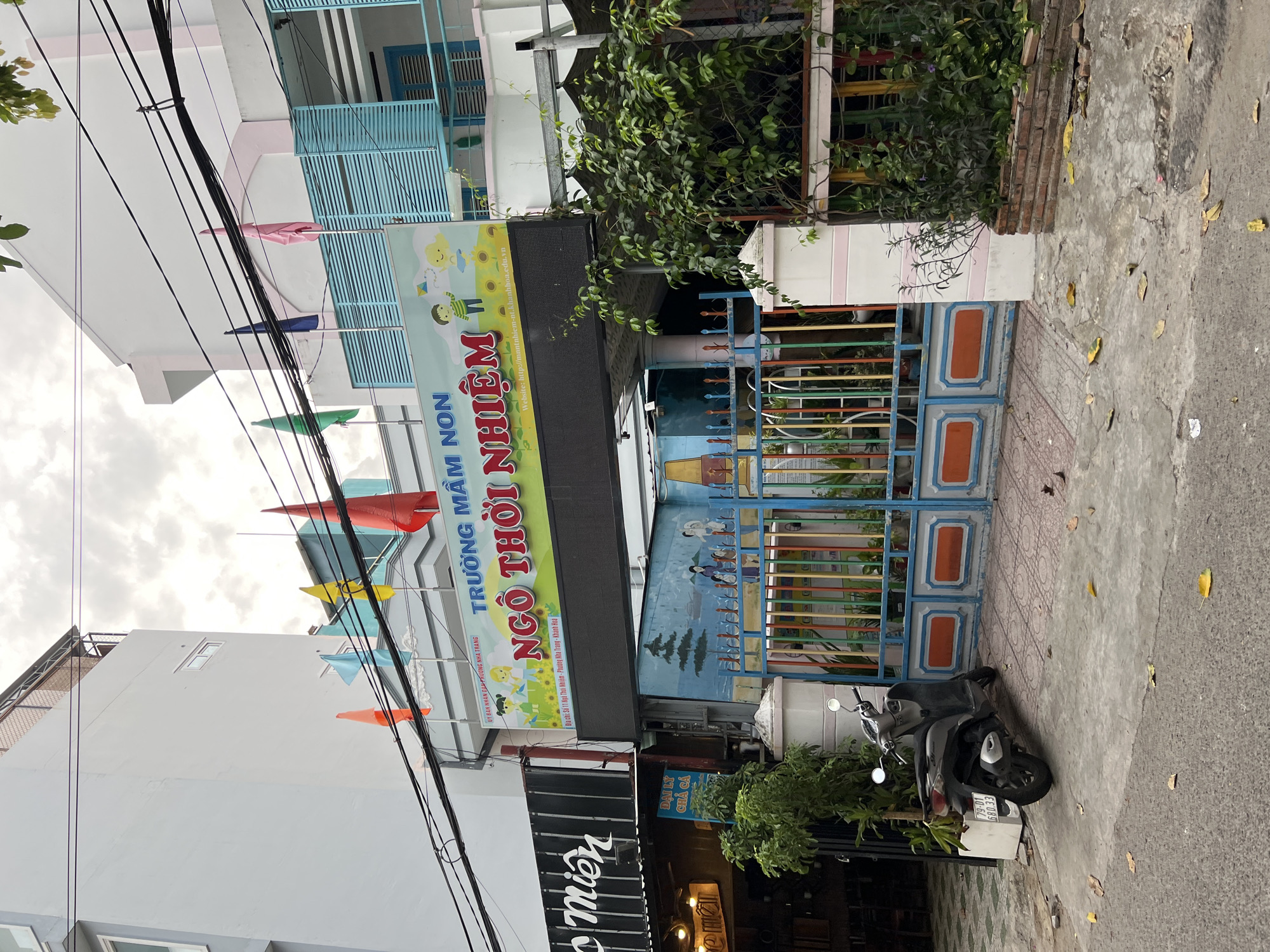 The image shows the entrance to a kindergarten in Nha Trang, Vietnam, called "Trường Mầm Non Ngô Thời Nhiệm". A large sign with colorful illustrations of children and a kite is displayed above the entrance. The building has a light blue facade with white trim, and a colorful gate with vertical bars in red, green, blue, and yellow. A motorbike is parked on the sidewalk in front of the gate. The street is paved and there are some fallen leaves on the ground. A mural depicting children and a landscape is visible on the wall behind the gate. The sky is overcast.