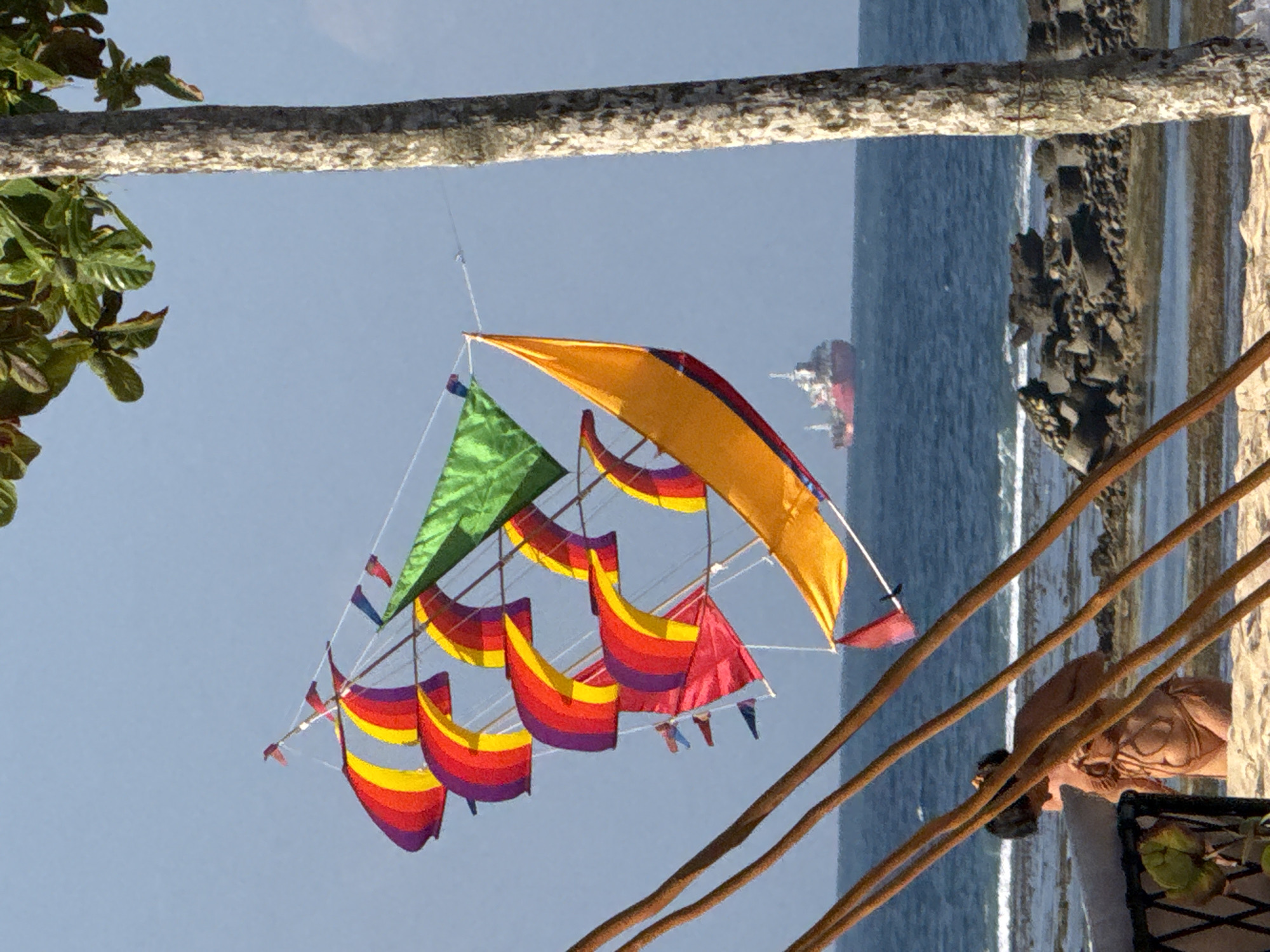 In Kuta Selatan, Indonesia, a large, multi-colored kite resembling a sailing ship is airborne against a clear sky. The kite features numerous sails in red, yellow, purple, and green, with a large yellow sail dominating the foreground. In the background, a red and white cargo ship is visible on the blue ocean. The foreground shows a sandy beach with weathered wooden poles, and a portion of a tree trunk with rough bark on the right. A person wearing a bikini is standing on the beach, facing away from the camera, with their back to the sea. Rocks and debris are scattered near the waterline.