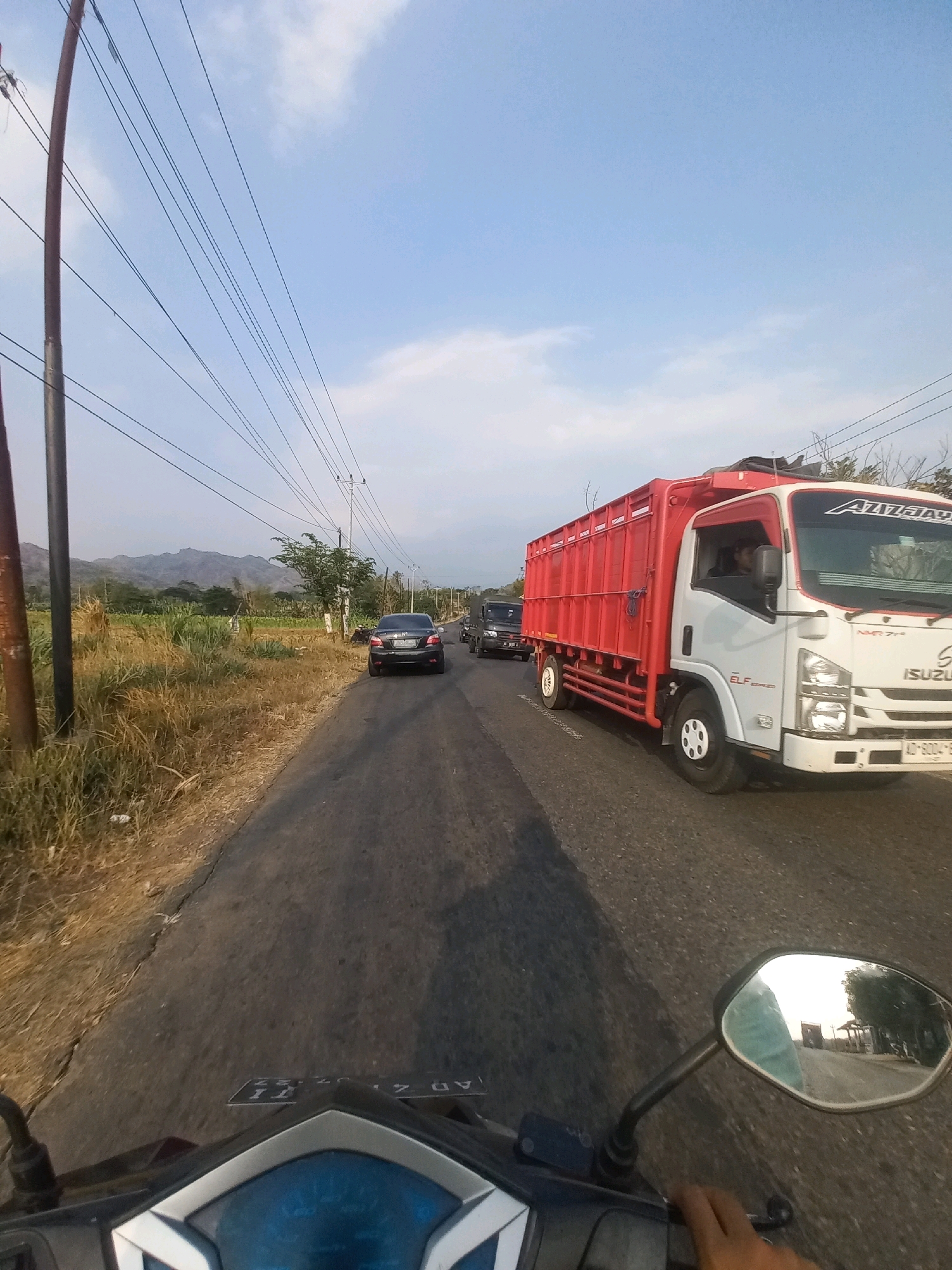 The image captures a street view in Baturetno, Indonesia, looking forward from a motorcycle. A paved road dominates the foreground, with a dry, grassy verge on the left and sparse vegetation on the right. In the distance, a truck with a red cargo container is visible in the right lane, and a dark sedan is in the left lane. Beyond the vehicles, a line of trees and a distant, hazy mountain range form the backdrop under a clear, pale blue sky with scattered clouds. Telephone poles and overhead wires are prominent in the sky. The motorcycle's dashboard and a handlebar mirror are visible in the bottom portion of the frame.