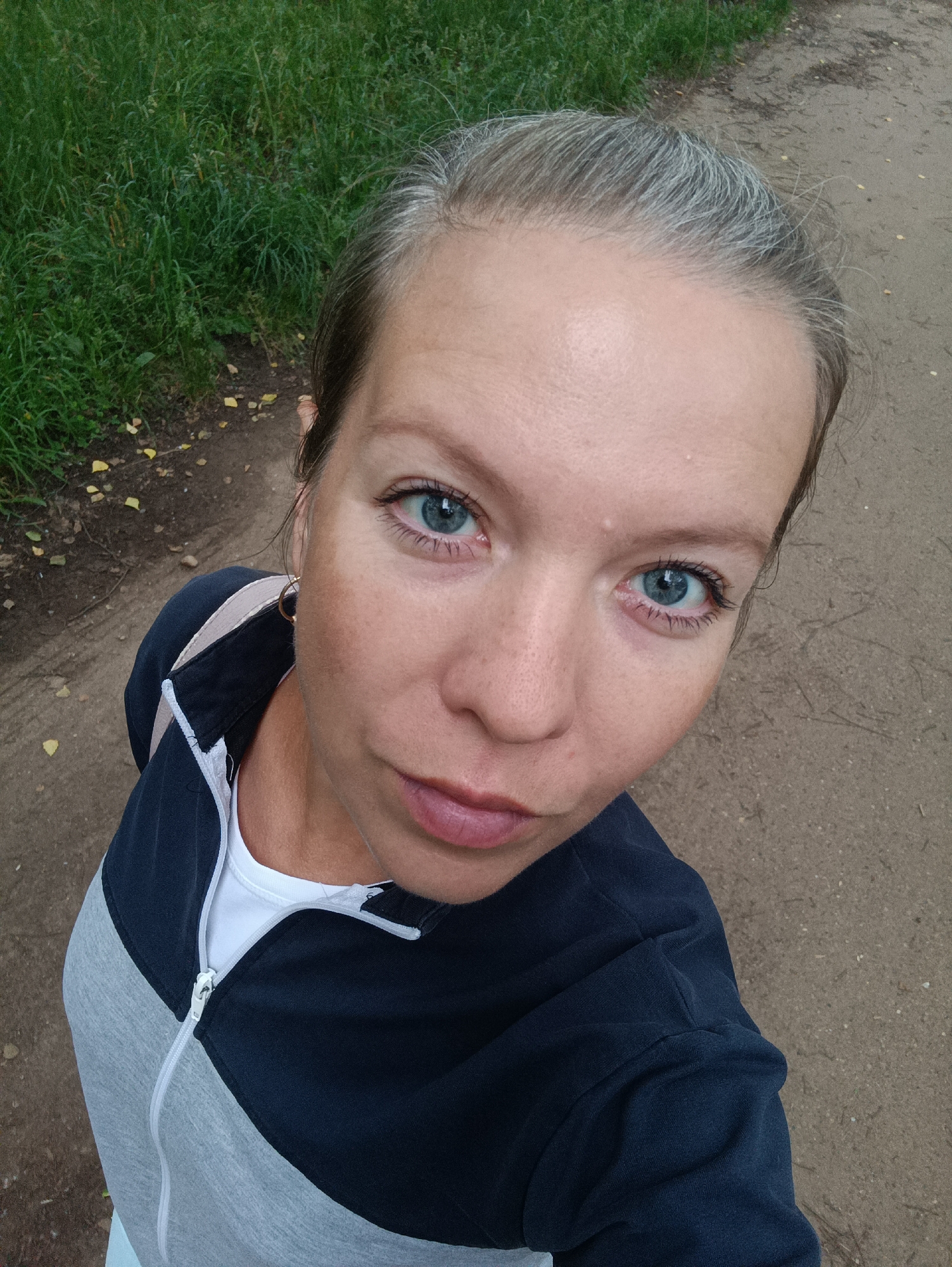 A high-angle, close-up shot features a woman with graying hair and blue eyes, looking directly at the camera. She is wearing a grey and dark blue zip-up jacket over a white shirt. Her lips are slightly parted. The background consists of a dirt path with sparse yellow leaves and green grass on the left side. The scene is likely outdoors in an area resembling a park or a walking trail within the city of Chaikovsky, Russia, given the path and vegetation.