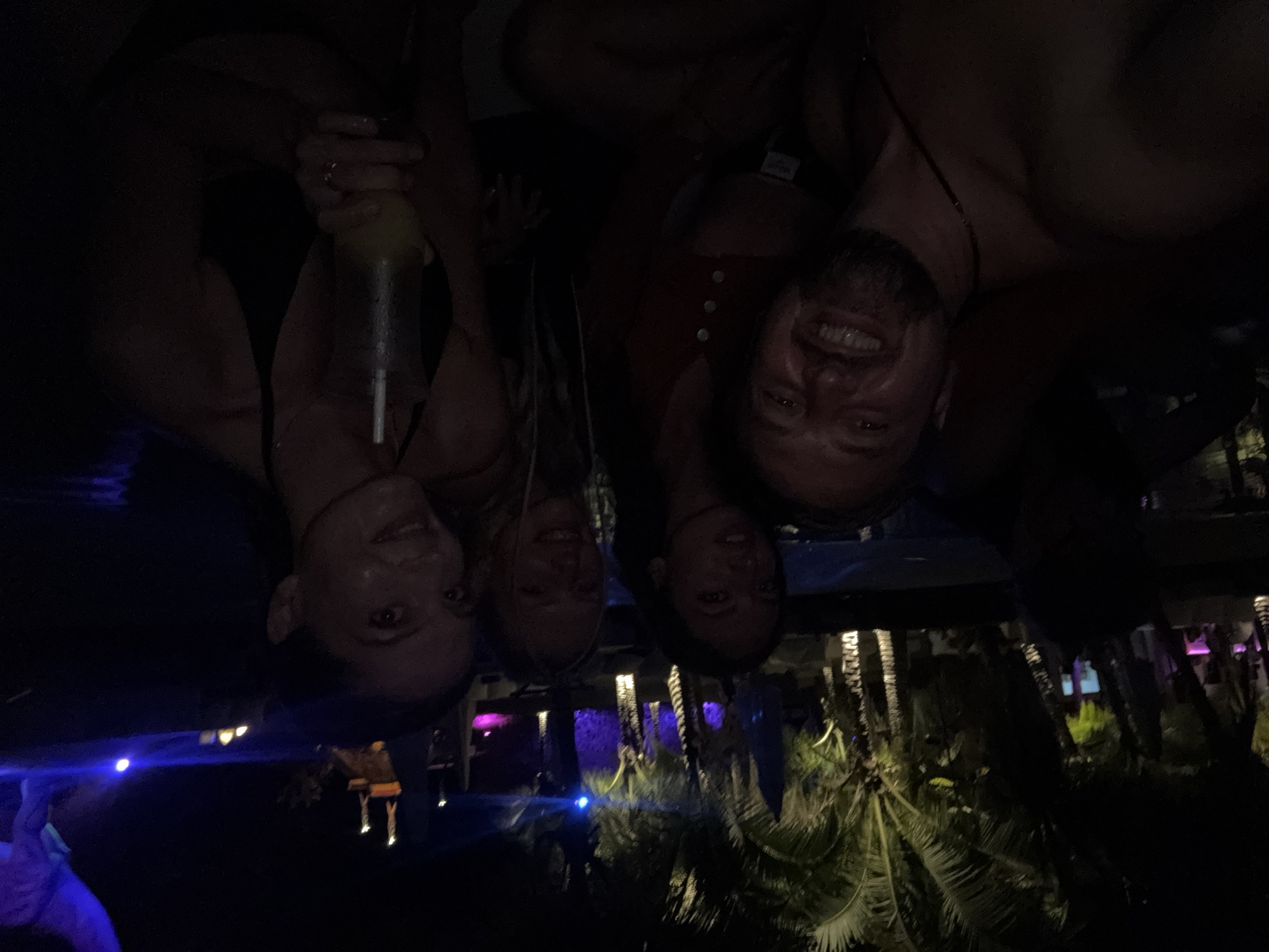 In this nighttime, outdoor image, four individuals are gathered, with three women and one man clearly visible. The man in the foreground, on the left, is smiling and looking towards the camera. To his right are two women, one of whom is holding a cocktail with a straw and a light-colored liquid. The third woman is partially obscured behind the other two. The background features palm trees illuminated by soft lighting, and in the distance, there are hints of structures and lights, suggesting an entertainment venue or resort. The overall ambiance is festive and social, with the individuals appearing to be enjoying themselves.