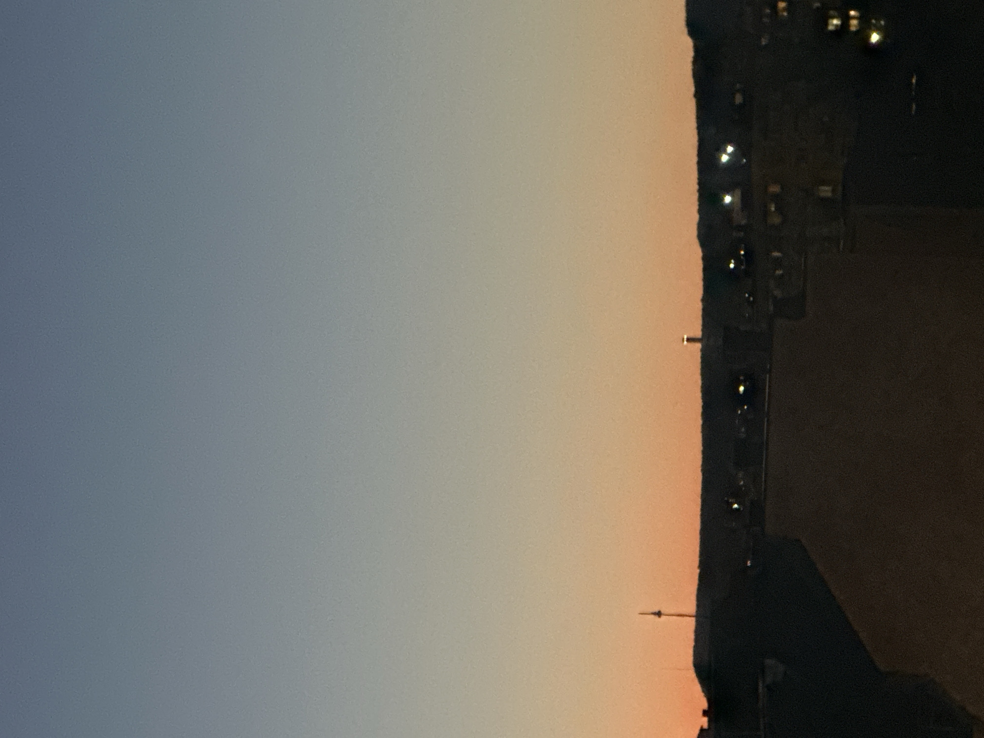 The image depicts a cityscape at dusk or dawn, likely Varna, Bulgaria, given the presence of the Asparuhov Bridge and the Varna TV Tower, which are prominent landmarks in the city. The sky transitions from a deep blue at the top to a warm orange near the horizon, indicating the time of day. Silhouetted against the sky are the outlines of buildings and hills. A tall, slender structure, consistent with the Varna TV Tower, is visible on the left side of the image. In the center, another illuminated structure, possibly part of a building or a smaller tower, stands out against the darker landscape. The lower portion of the image shows darker, indistinct shapes of buildings, with some lights visible, suggesting the city is beginning to illuminate as night falls or the day breaks. The overall atmosphere is serene and quiet, capturing a transitional moment in the urban environment.