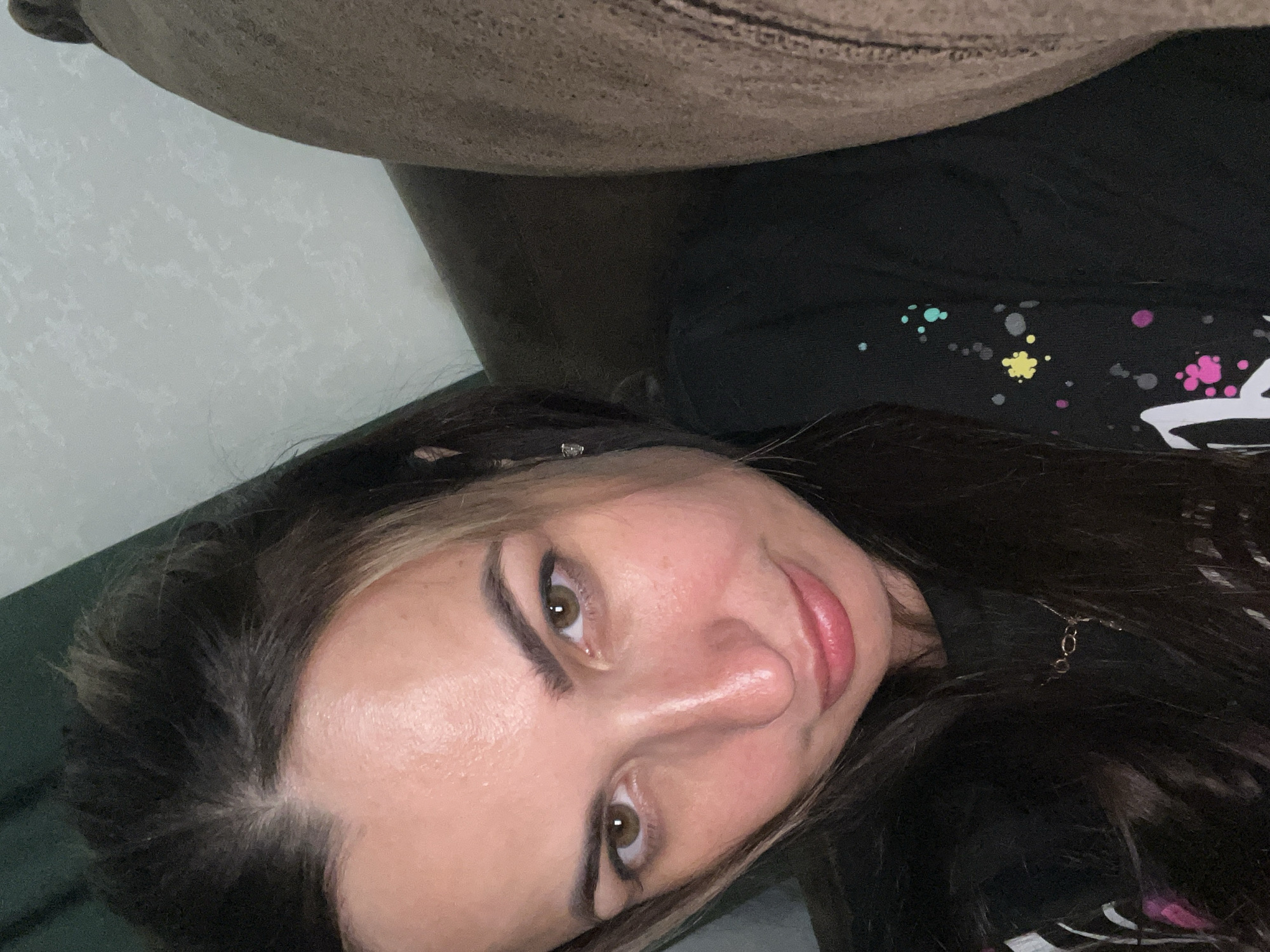 A close-up selfie captures a woman with long, dark hair with blonde highlights, looking directly at the camera. She has brown eyes, prominent eyebrows, and fair skin with a touch of pink on her cheeks. Her lips are slightly parted, and she's wearing a black t-shirt with a colorful paint-splatter design and a delicate necklace. She is leaning against a brown sofa, with a textured wall and a dark green curtain visible in the background. The lighting is soft, casting a gentle glow on her face.