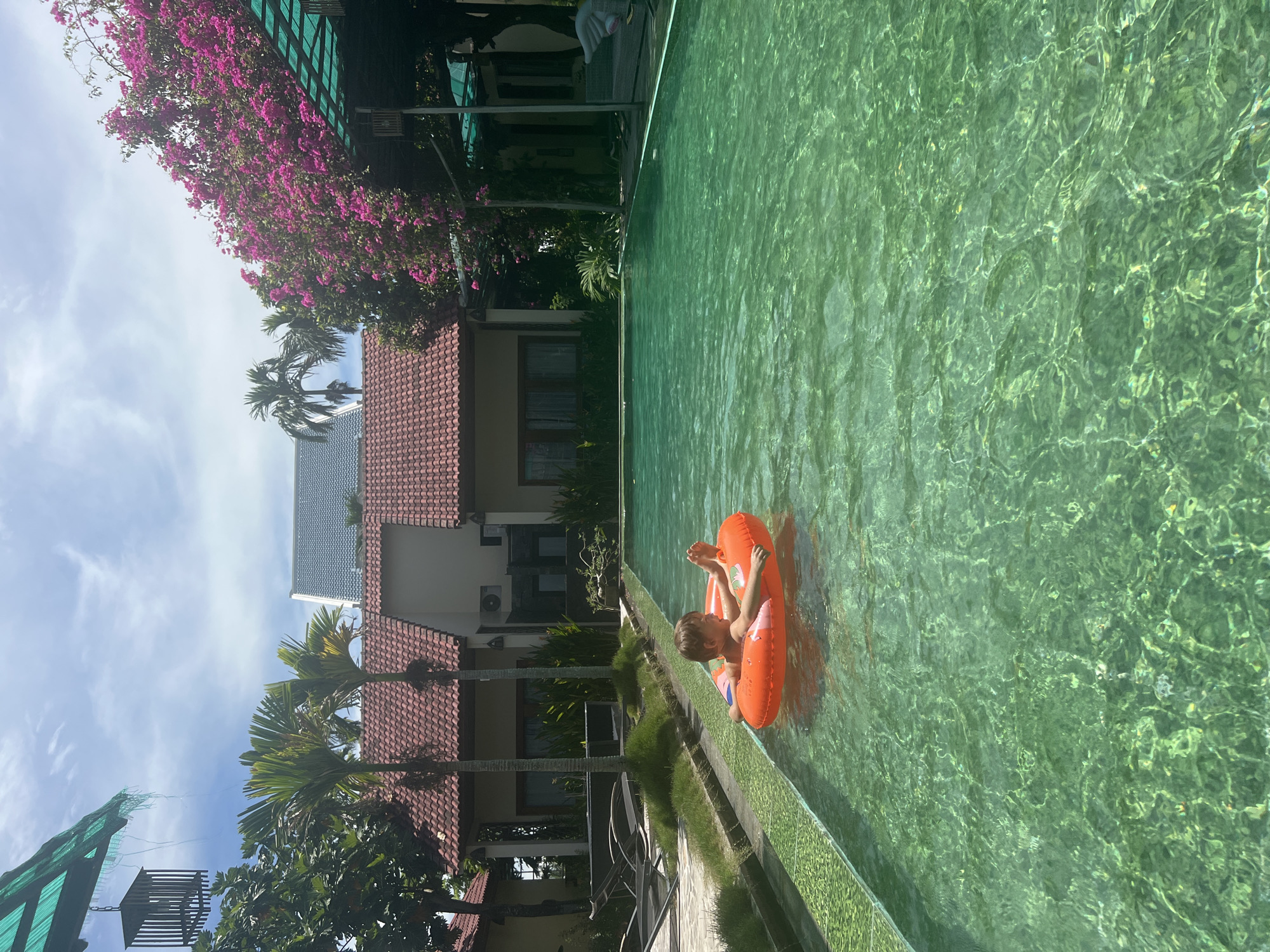 A young boy floats in a swimming pool on an orange inner tube. He is in the center of the frame, facing forward with his arms raised. The pool water is clear and green, with sunlight glinting off the surface. Behind the pool, there are several buildings with red tiled roofs and palm trees. Pink bougainvillea flowers bloom on a trellis to the right. The scene appears to be a resort in Hoi An, Vietnam, given the architectural style and lush vegetation.